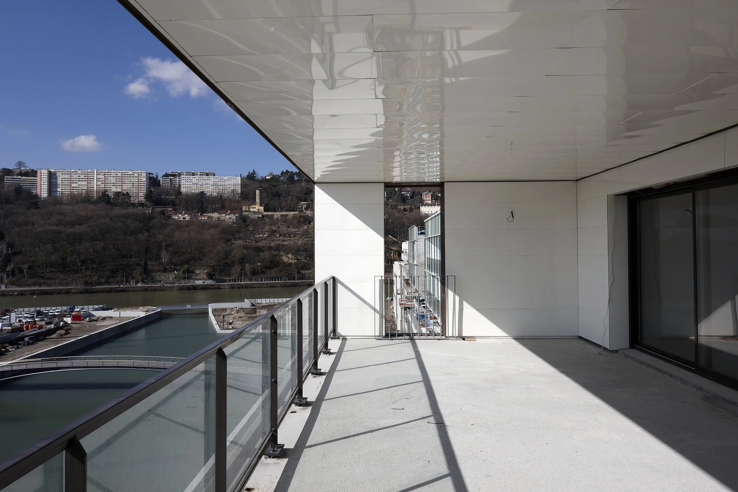 Vue d'une terrasse d'un bâtiment moderne avec garde-corps en verre, surplombant une rivière ou un canal, avec des bâtiments résidentiels en arrière-plan, par un jour ensoleillé avec un ciel bleu et quelques nuages.