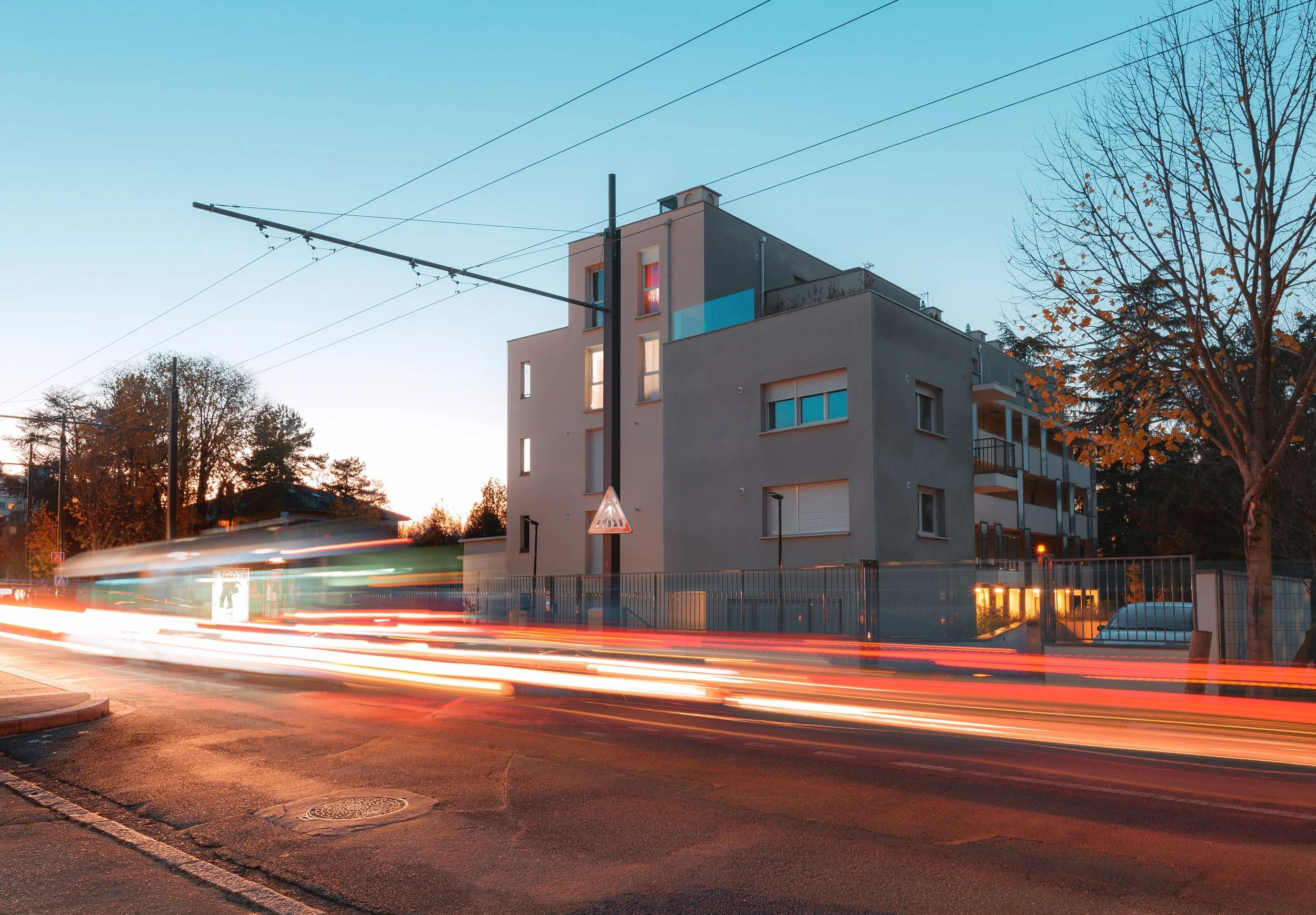 Une vue urbaine au coucher du soleil avec un bâtiment résidentiel moderne, un arbre aux feuilles d'automne, et des traînées lumineuses de voitures en mouvement sur la rue.