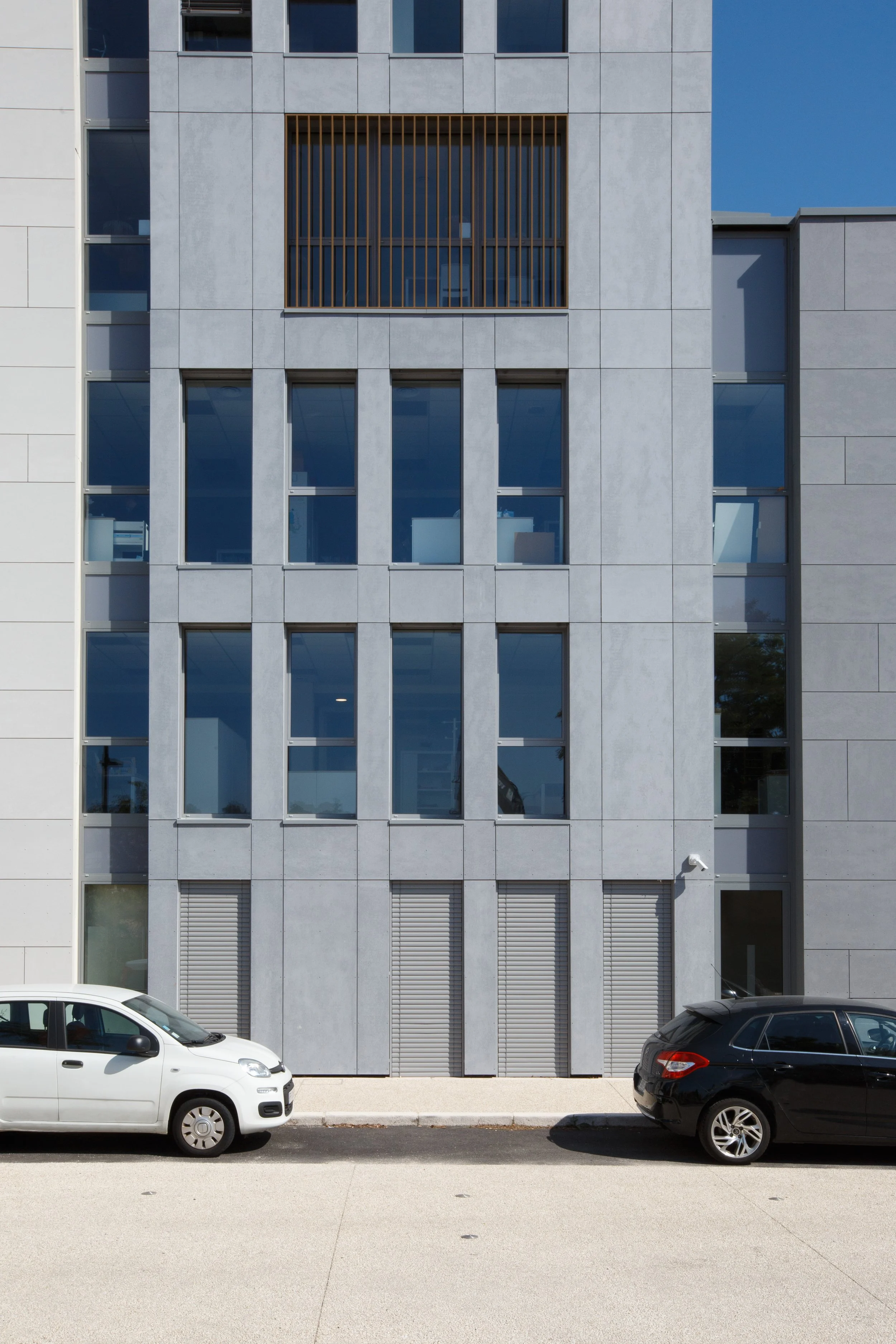 Façade d'un bâtiment moderne avec fenêtres rectangulaires et lamelles en bois, deux voitures garées devant et un ciel bleu en arrière-plan.