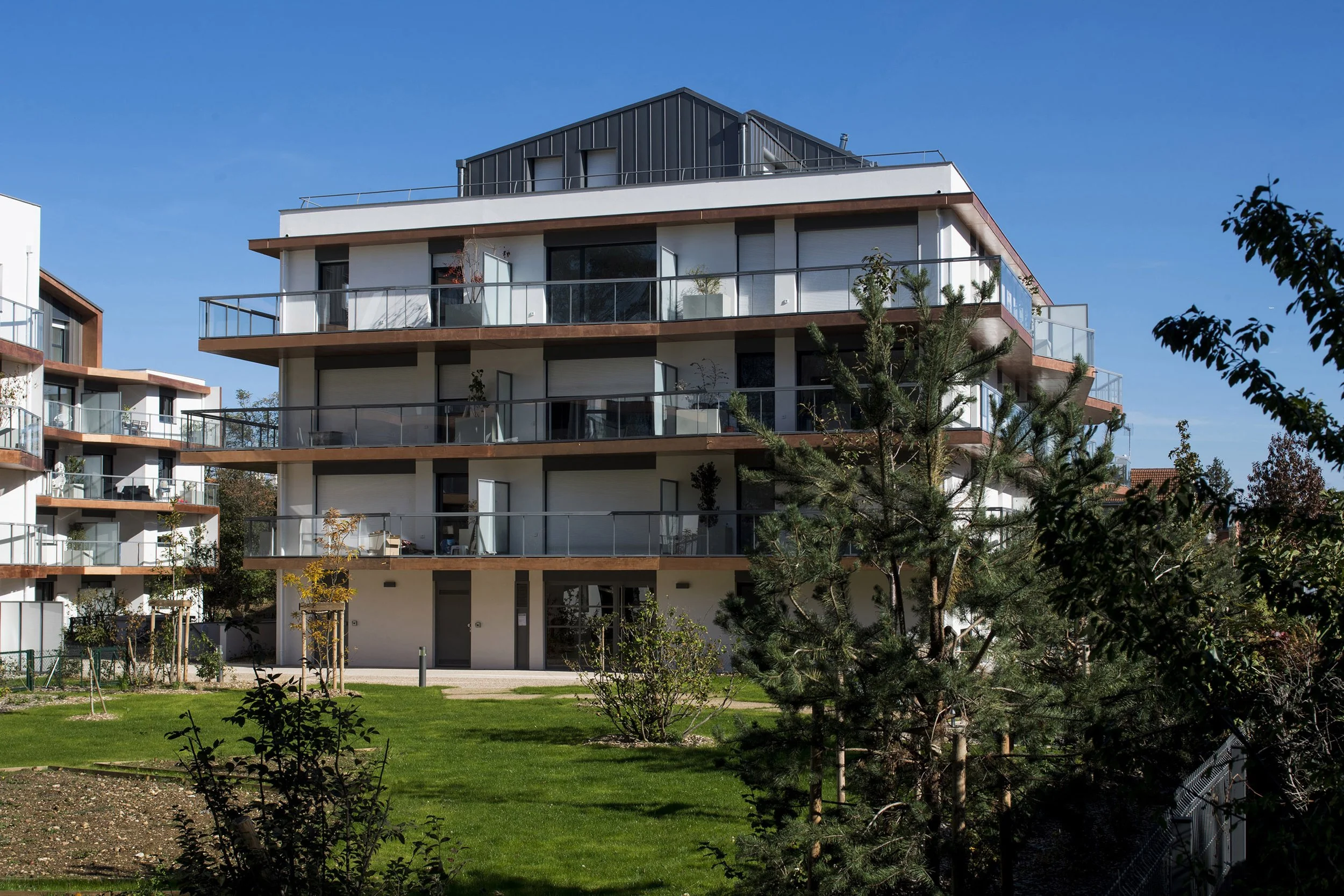 Appartement moderne à plusieurs étages avec balcons et terrasses, entouré d'espaces verts et d'arbres, sous un ciel bleu clair.