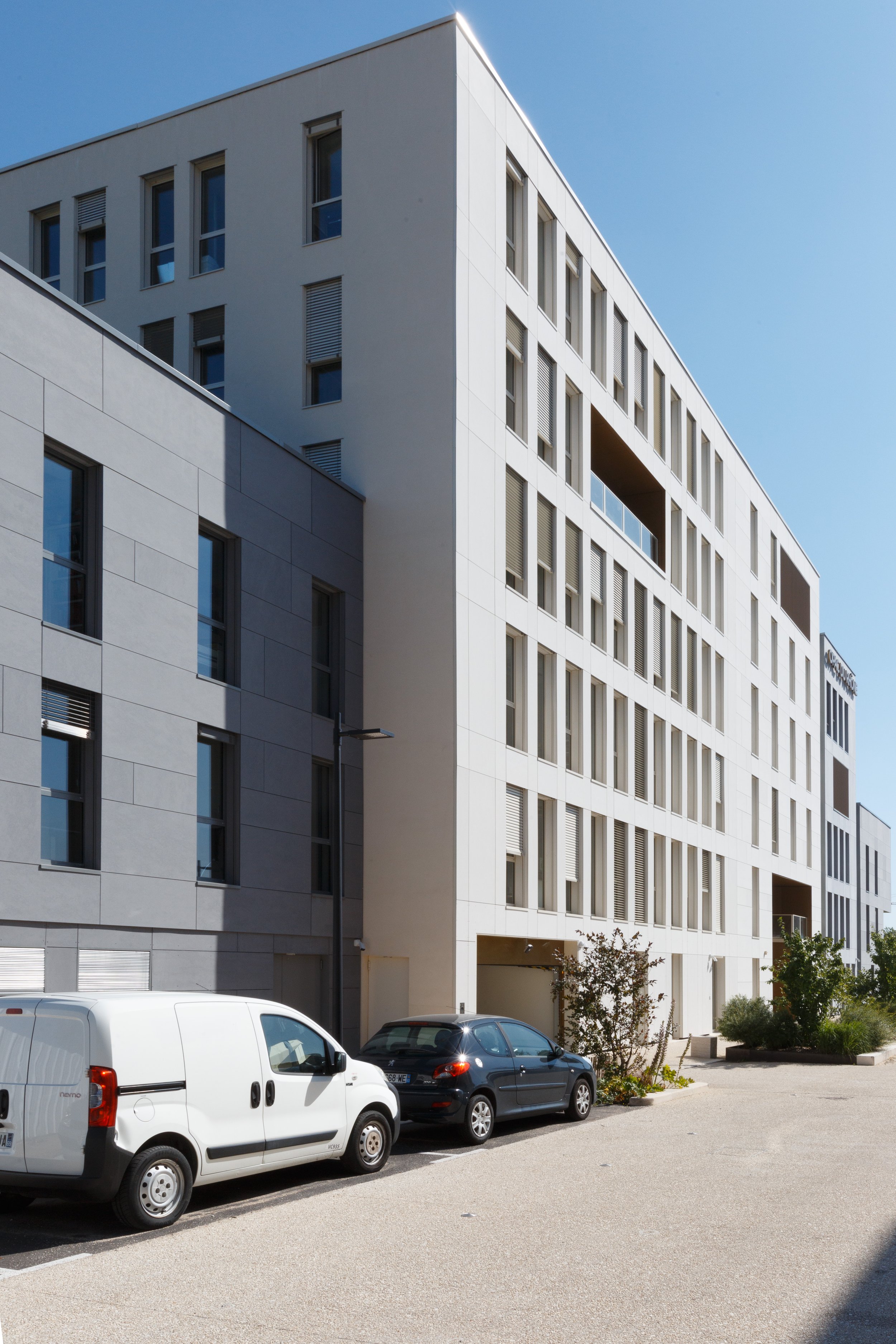 Bâtiment moderne blanc avec plusieurs fenêtres, stationnement avec voitures, arbres et un lampadaire dans un environnement urbain sous un ciel clair.