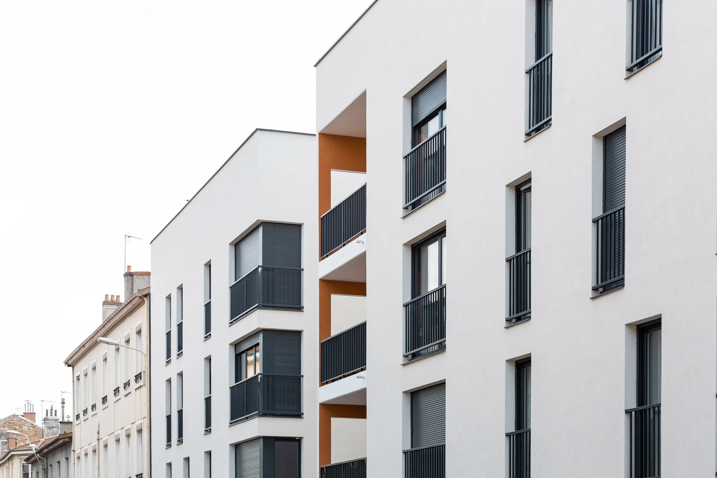 Immeuble moderne avec plusieurs balcons en fer noir, fenêtres à volets roulants, structure blanche avec accents beige, vue de près avec des bâtiments traditionnels en arrière-plan.