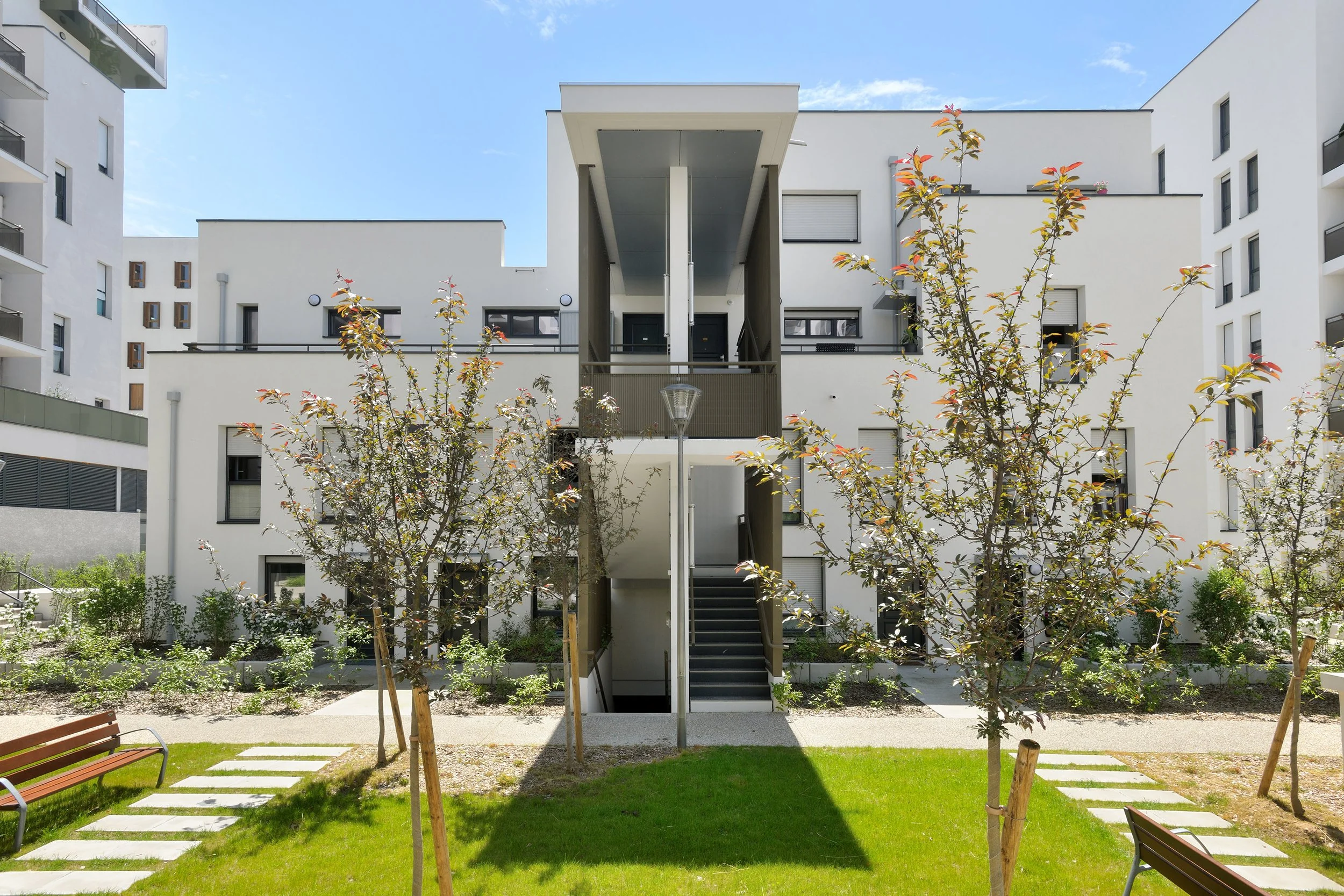 Appartement moderne blanc avec balcon, escalier extérieur et arbres dans la cour, ciel bleu et nuages à l'arrière-plan.