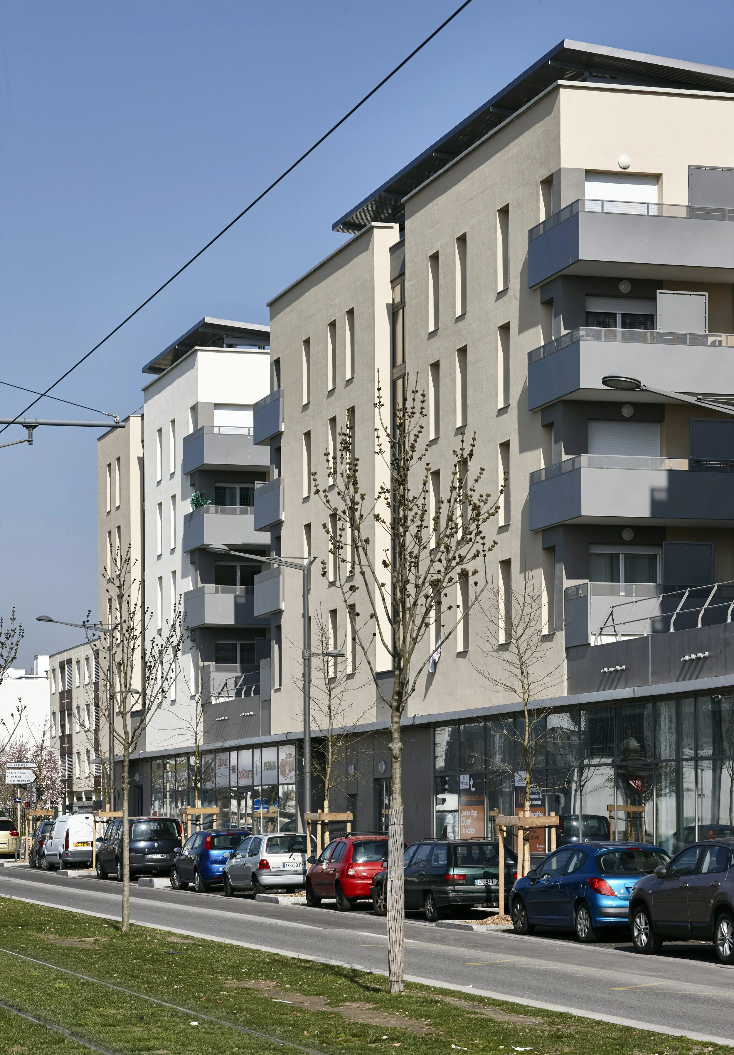 Bâtiment moderne avec plusieurs étages et balcons, stationnement avec des voitures, trottoir et arbres en premier plan, ciel clair et ensoleillé.
