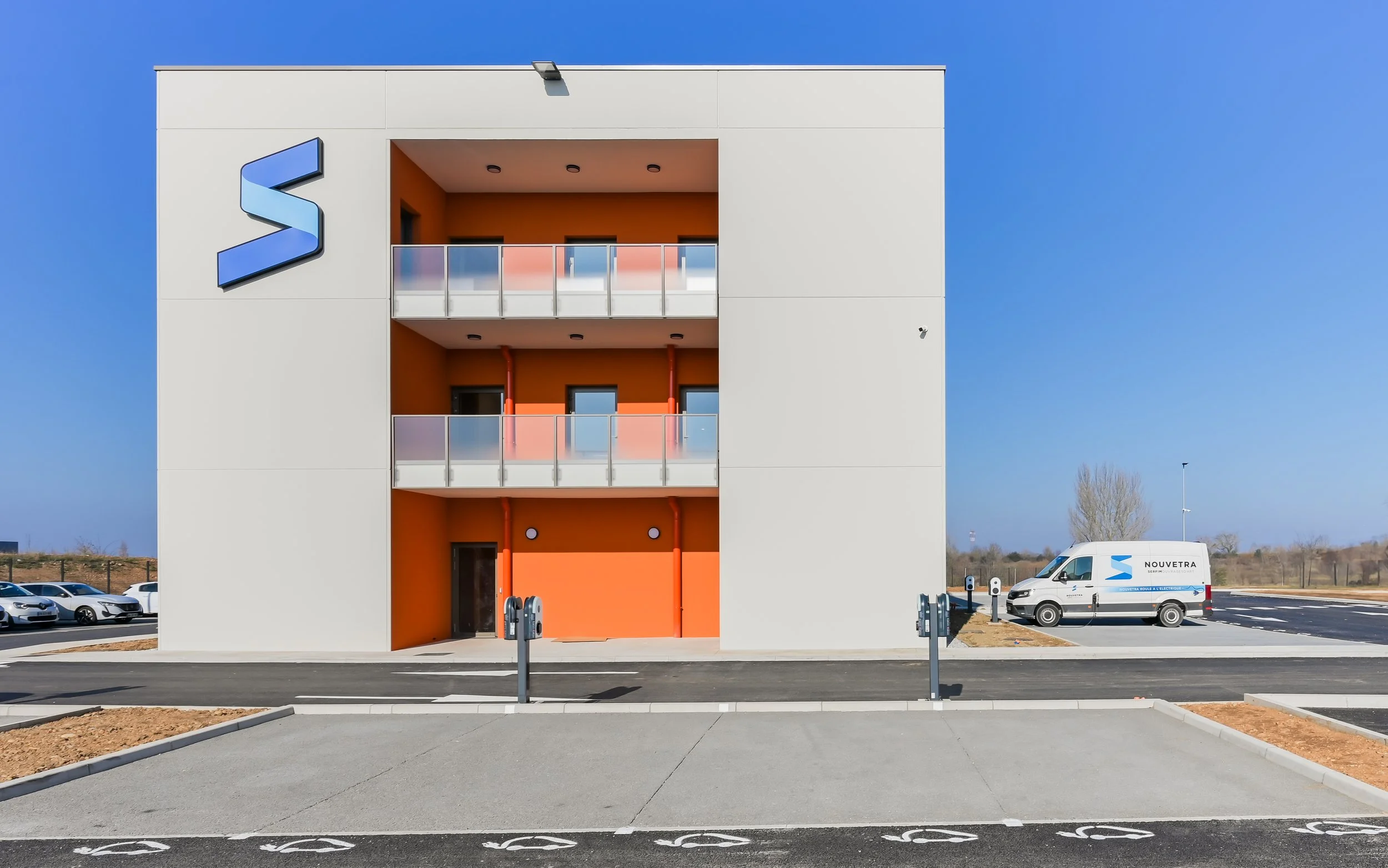 Bâtiment moderne avec trois balcons, signe en forme de S sur la facade, parking avec voitures et camion de livraison électrique, ciel bleu clair.