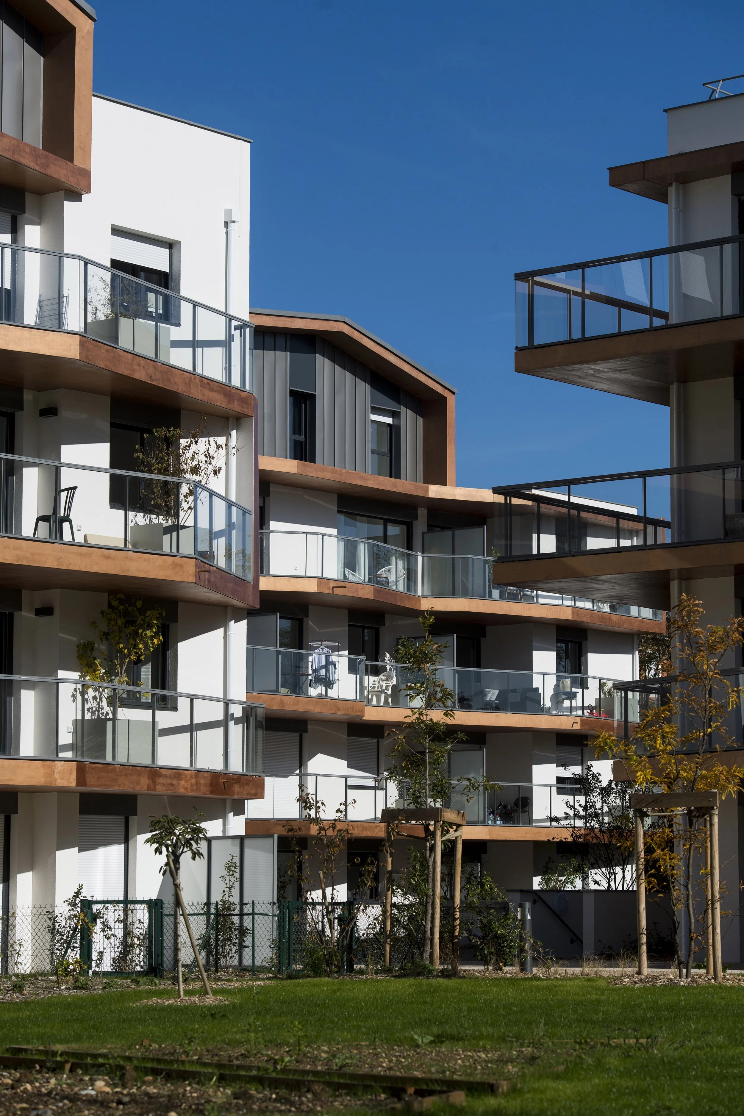 Appartement moderne avec plusieurs étages, balcons en verre, façade mixte blanche et gris foncé, sous un ciel bleu clair, avec un espace vert et des jeunes arbres en premier plan.