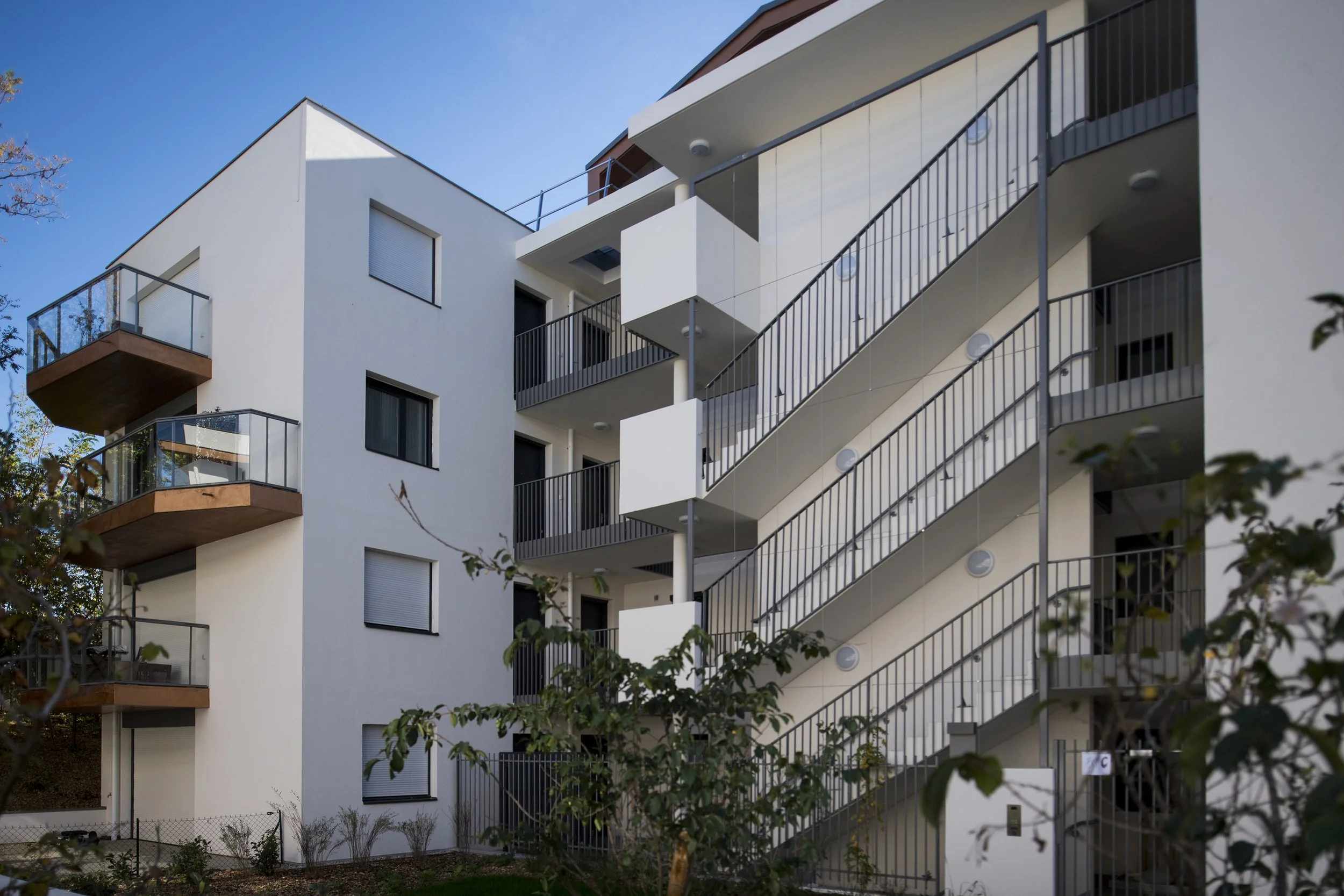 Appartement moderne à plusieurs étages avec balcons en verre et une rampe d'escalier extérieure en métal, sous un ciel bleu.