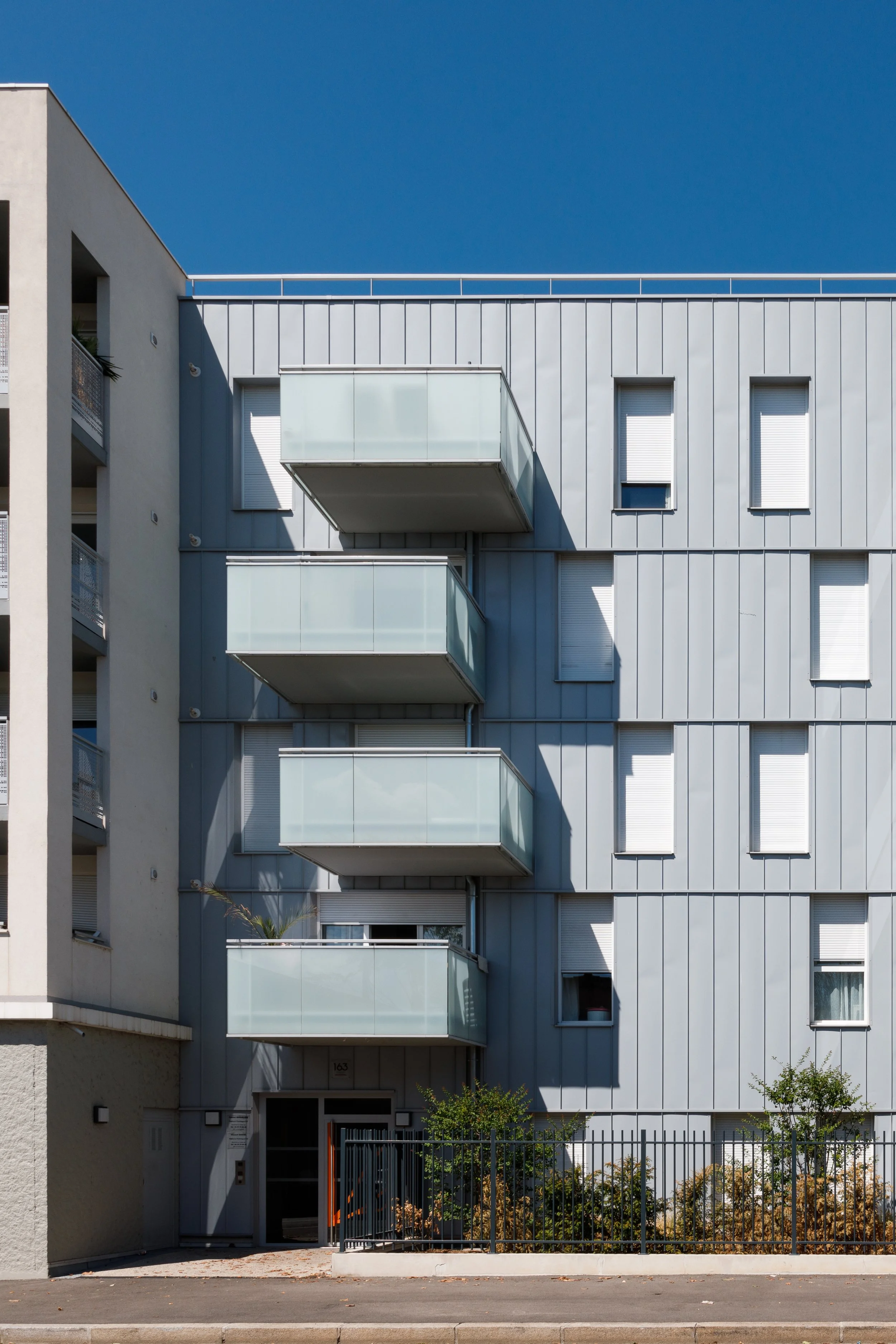 Immeuble résidentiel moderne avec balcons en verre, fenêtres à volets roulants, et petite végétation à l'avant, sous un ciel bleu clair.