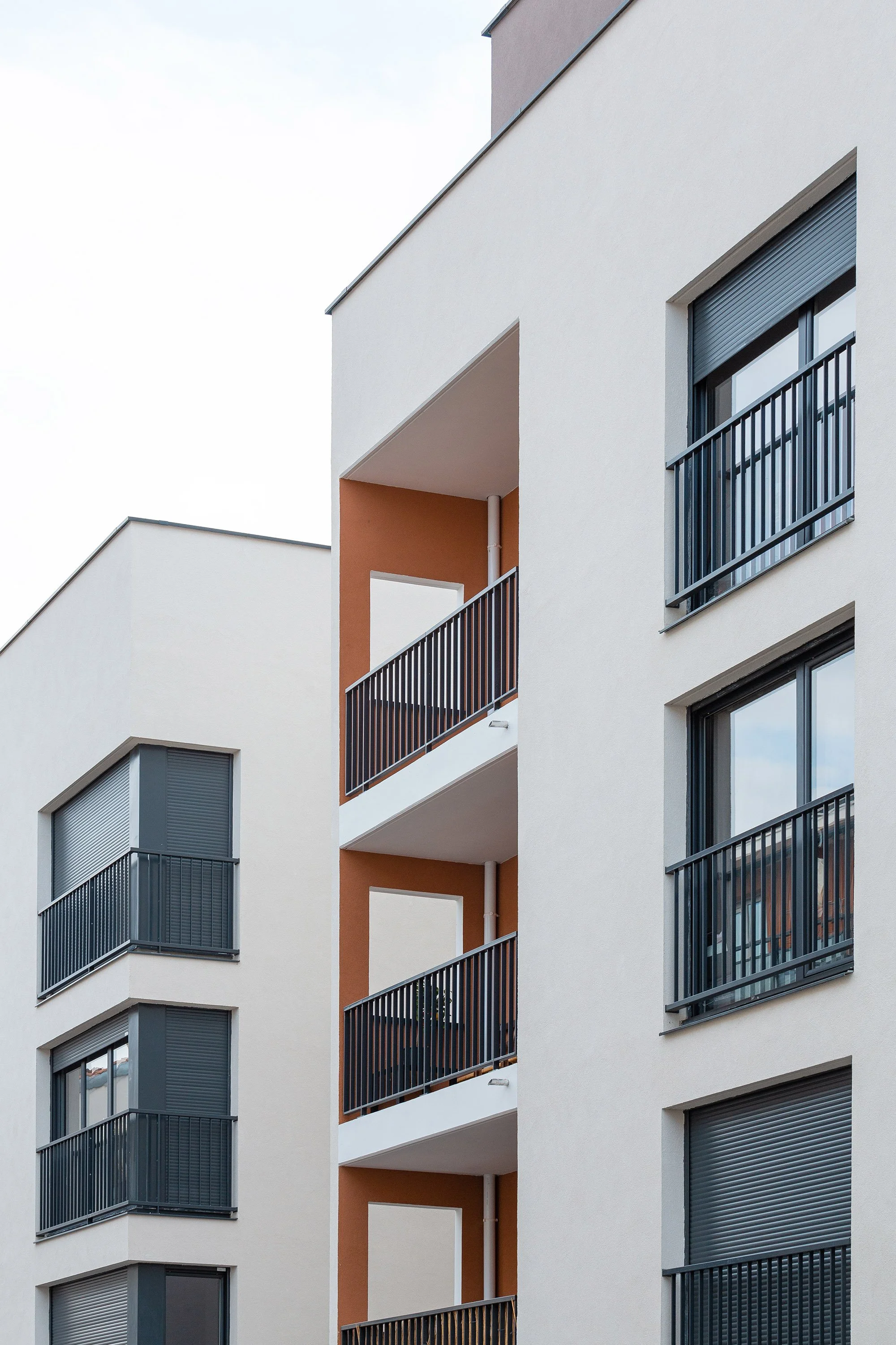 Vue d'un immeuble moderne avec plusieurs balcons, fenêtres avec volets roulants gris et murs blanc-beige, sous un ciel clair.