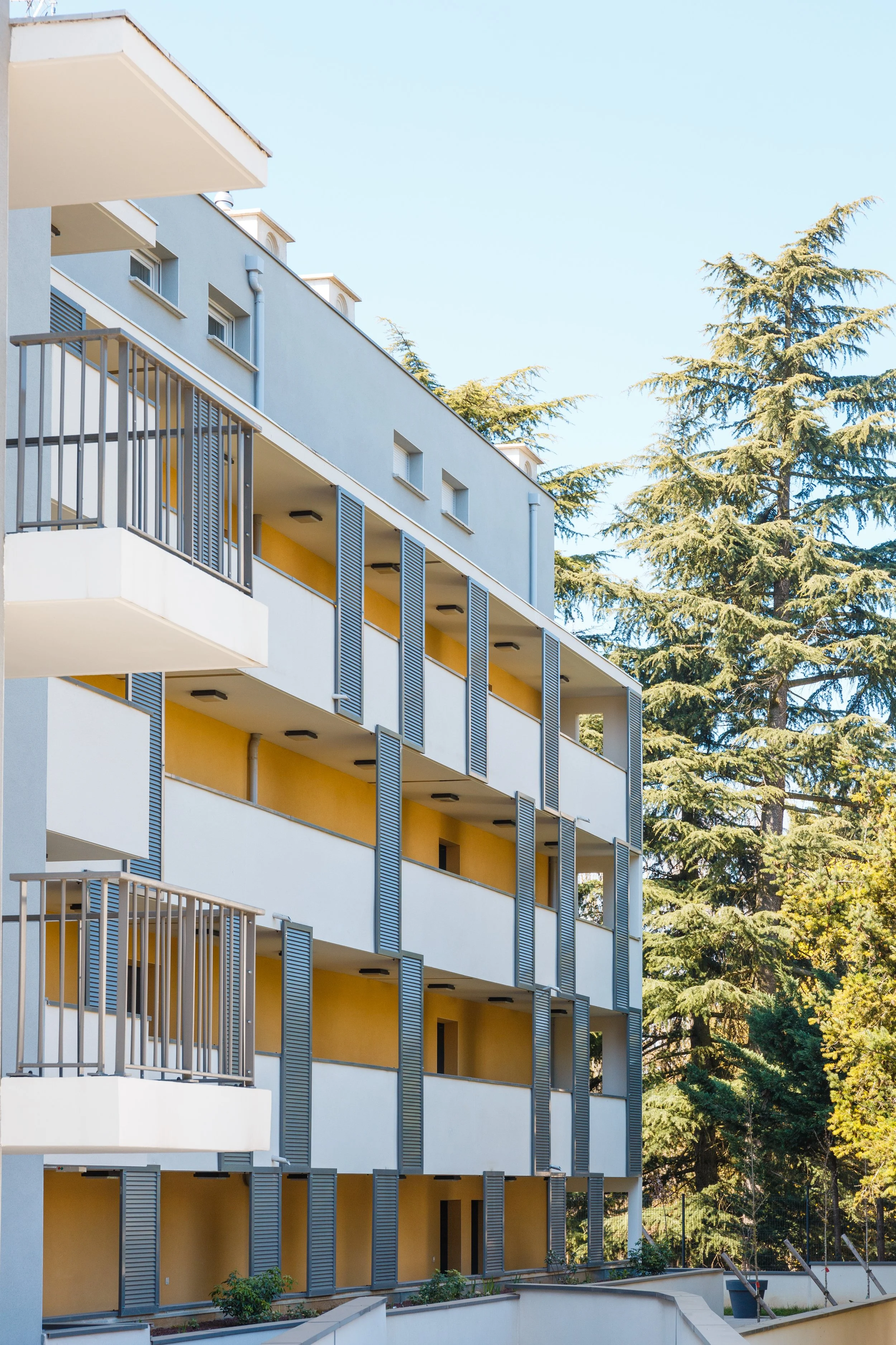 Immeuble résidentiel moderne avec plusieurs balcons et fenêtres, entouré d'arbres en plein air, sous un ciel clair.