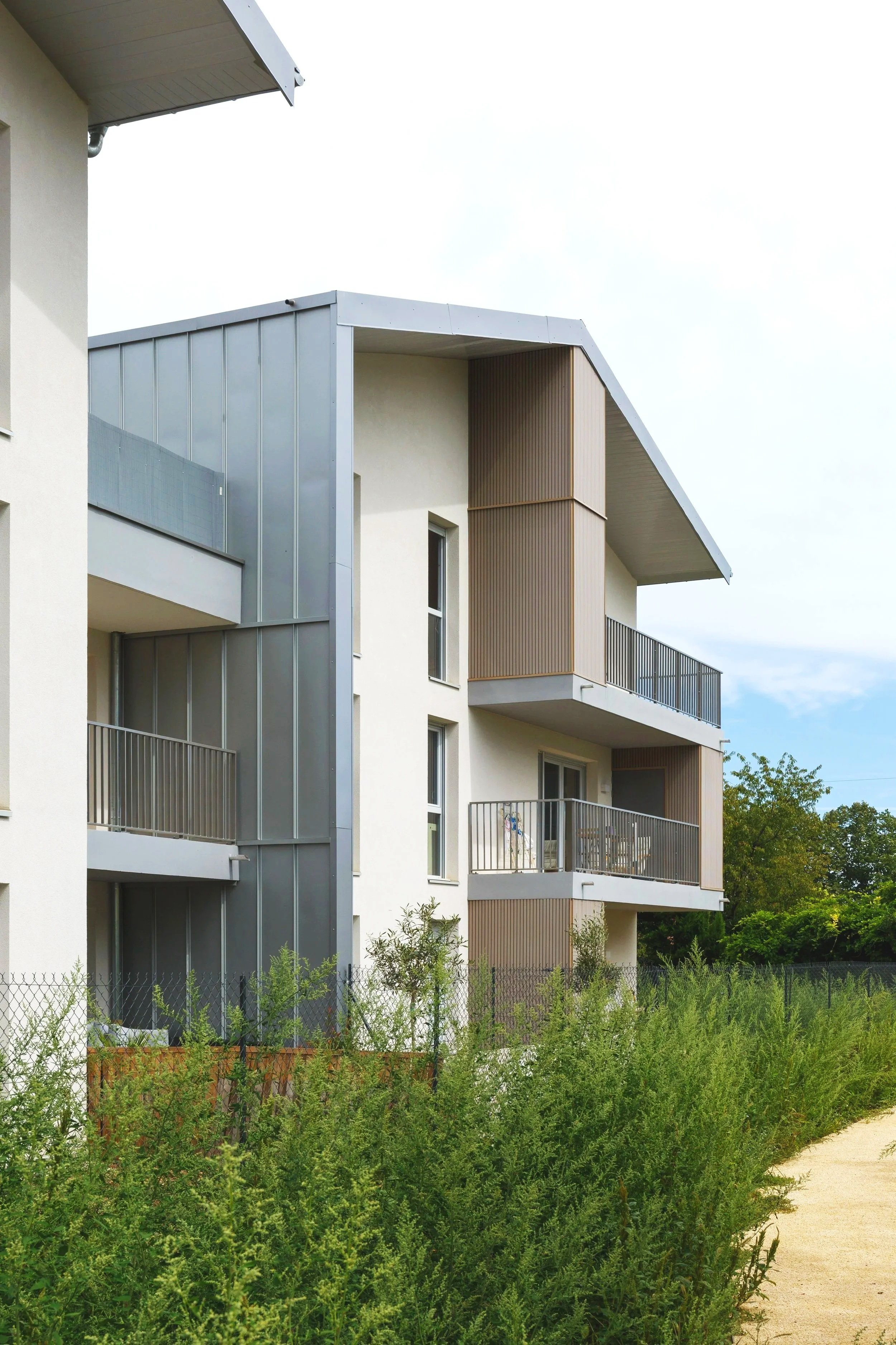 Un immeuble résidentiel moderne avec plusieurs balcons en béton, façade en apparence en béton clair, avec des éléments en bois et en métal, entouré de végétation luxuriante et d'un chemin de terre.