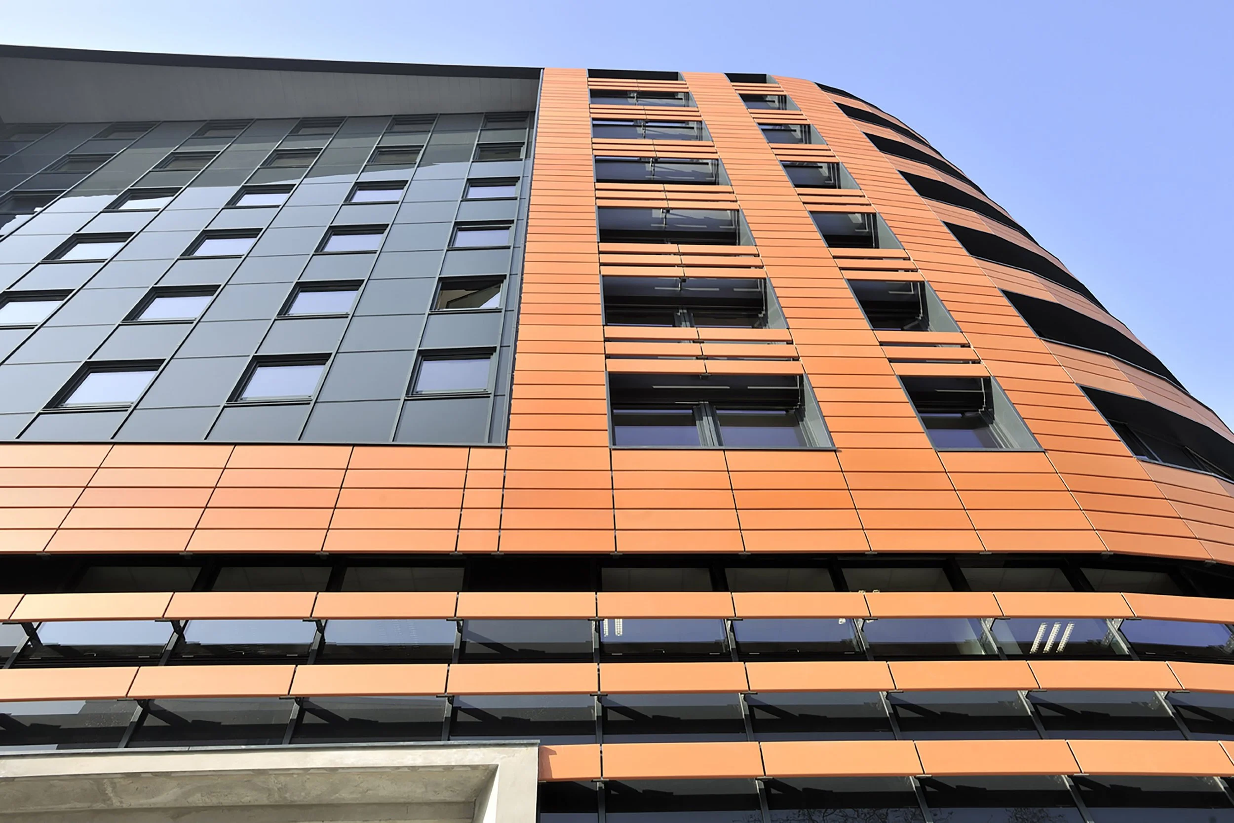 Facade moderne d'un bâtiment avec des fenêtres rectangulaires et des panneaux en métal orange et gris, vue en contre-plongée contre un ciel clair.