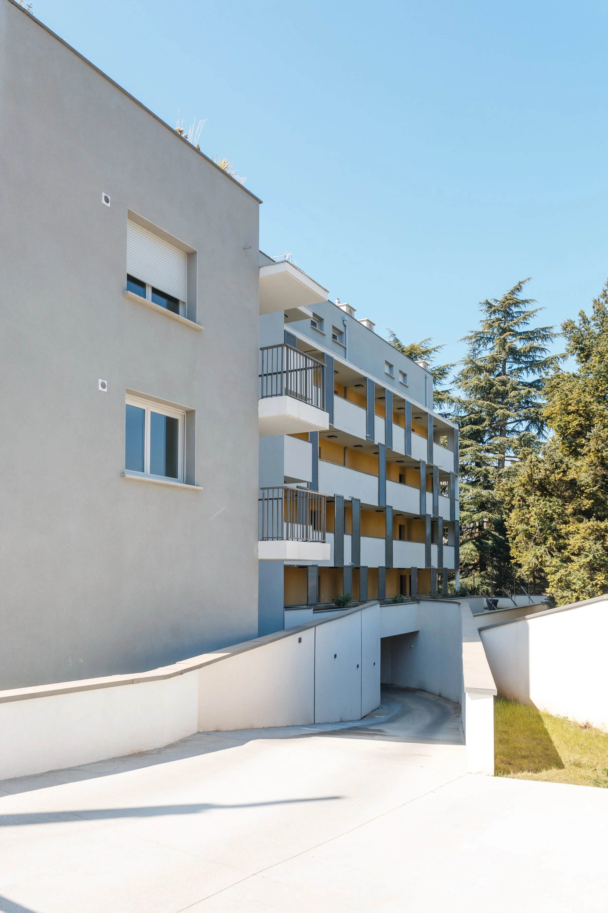 Une vue d'un bâtiment résidentiel moderne avec plusieurs balcons, fenêtres à volets et une façade blanche, sur fond d'arbres et un ciel bleu clair.