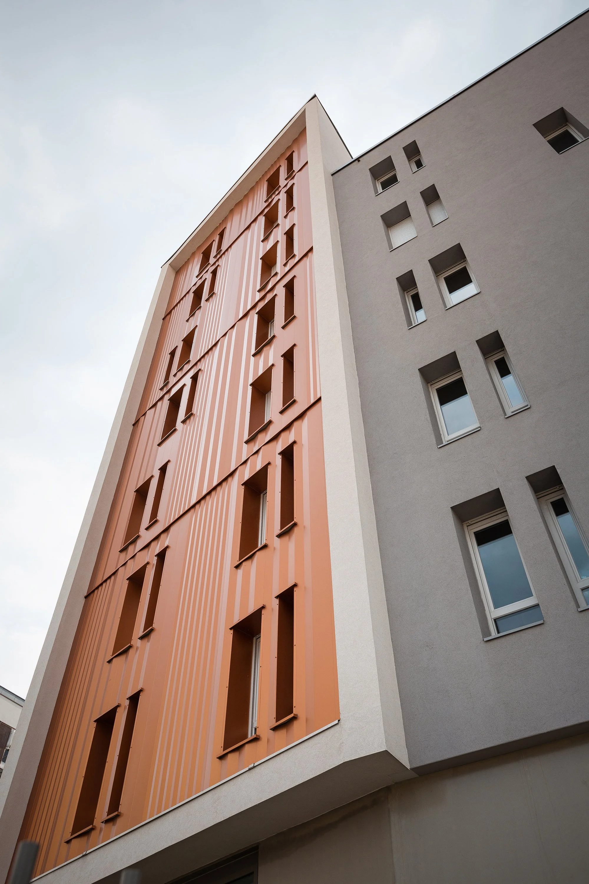 Une photographie d'un immeuble moderne avec une façade orange et une autre grise, vue en contre-plongée, sous un ciel nuageux.
