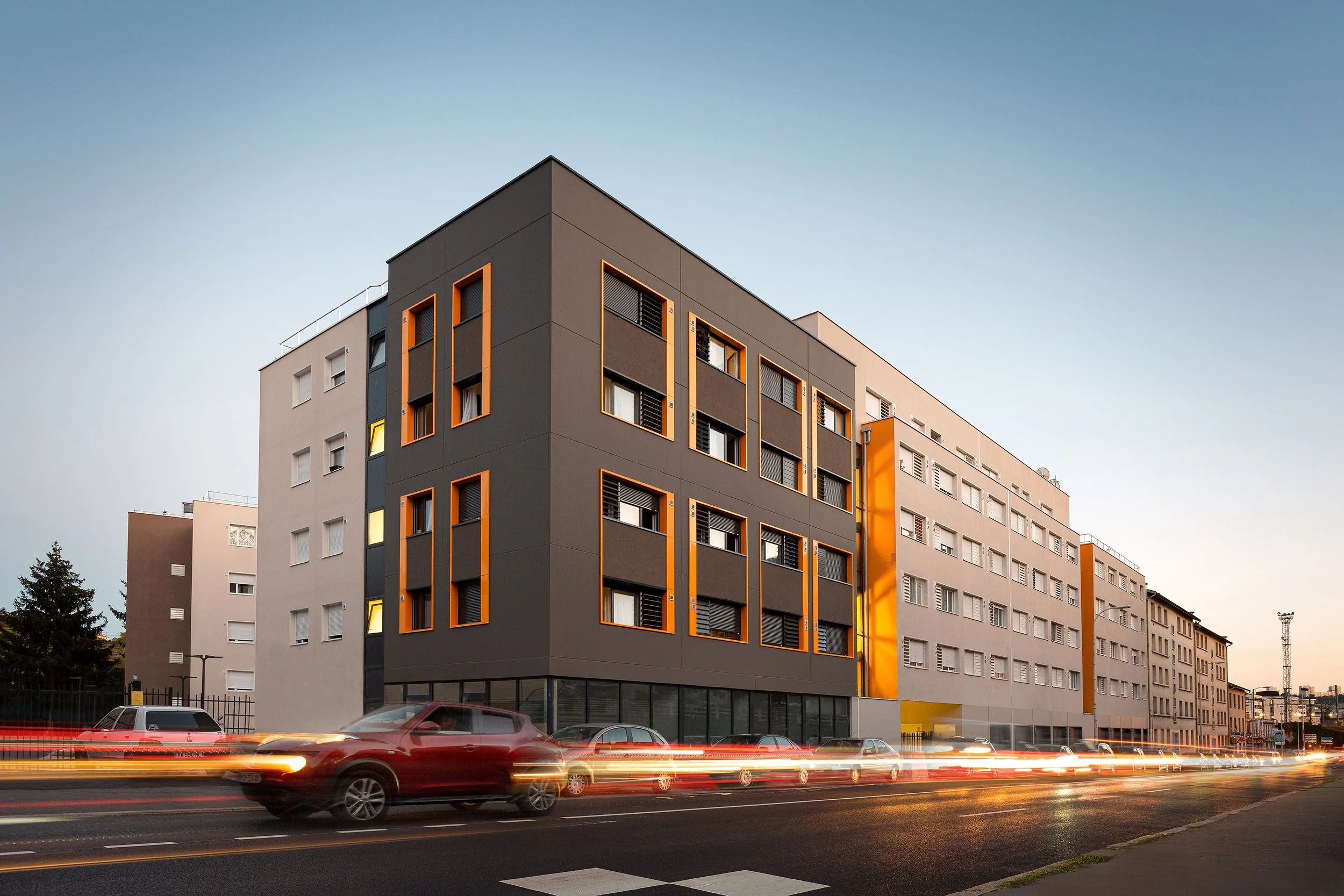 Immeuble résidentiel moderne avec façade grise et oranges, grandes fenêtres rectangulaires, voitures en mouvement au premier plan, ciel clair au crépuscule.