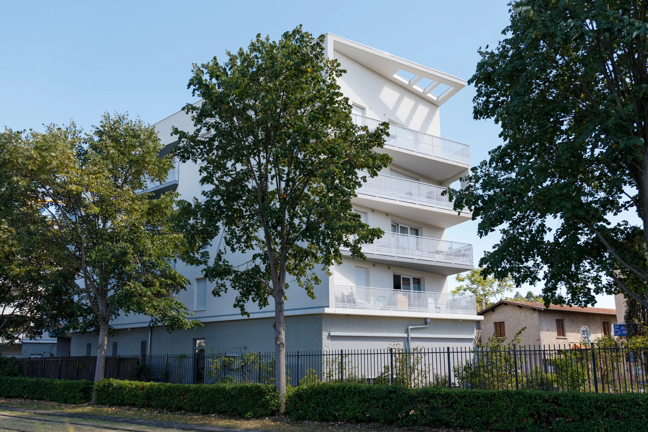 Un immeuble résidentiel blanc avec de multiples balcons, entouré d'arbres verts, sous un ciel clair.