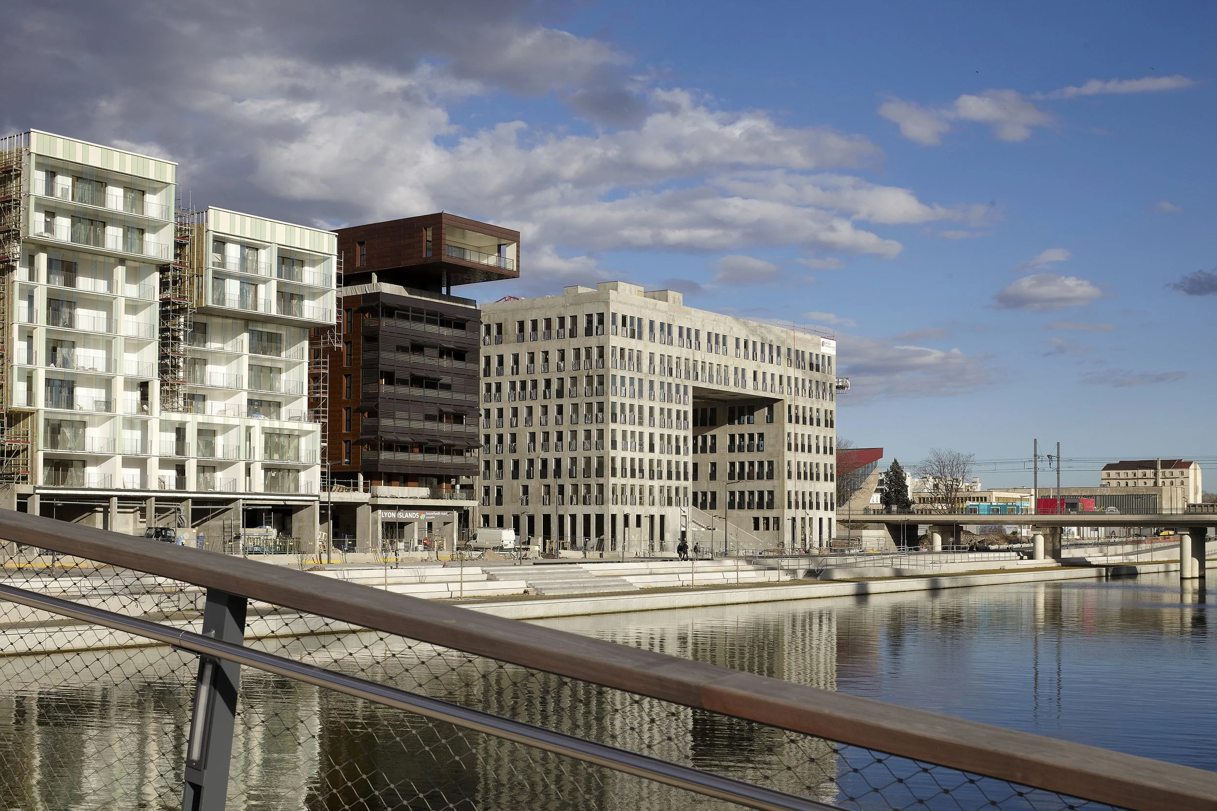 Bâtiments modernes en construction à côté d'une rivière, avec un pont piétonnier en premier plan et un ciel partiellement nuageux.