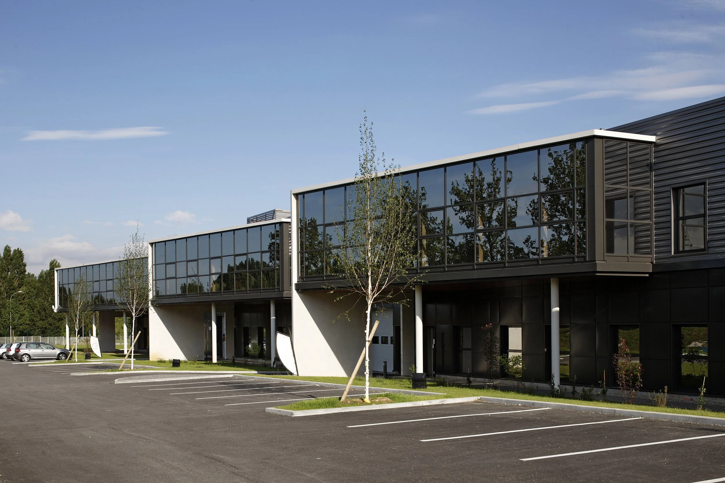 Bâtiment moderne avec de grandes fenêtres en verre, parking vide, jeunes arbres plantés autour, ciel bleu avec quelques nuages.