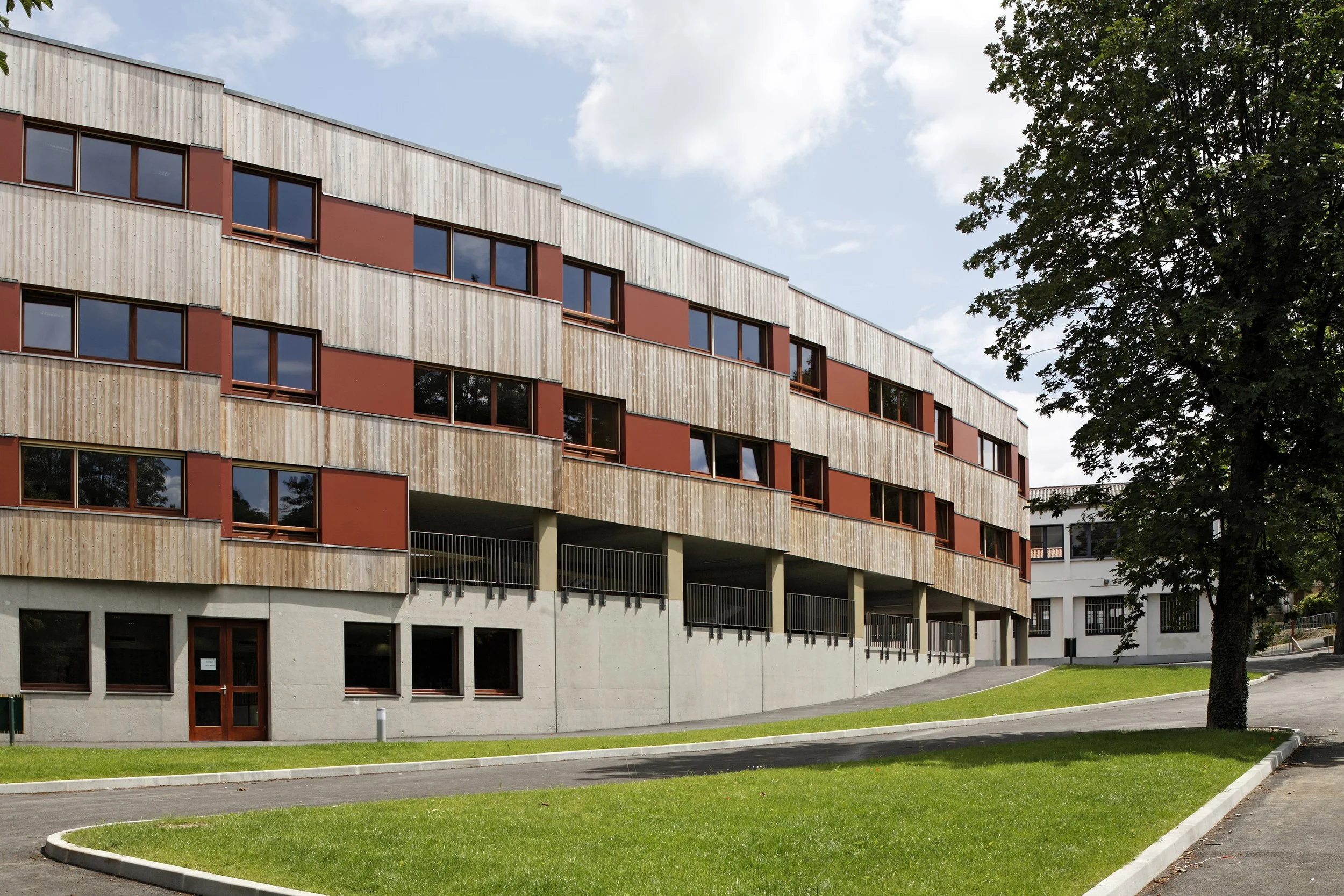 Bâtiment moderne en pierre avec fenêtres en bois et un arbre à côté, sous un ciel partiellement nuageux.