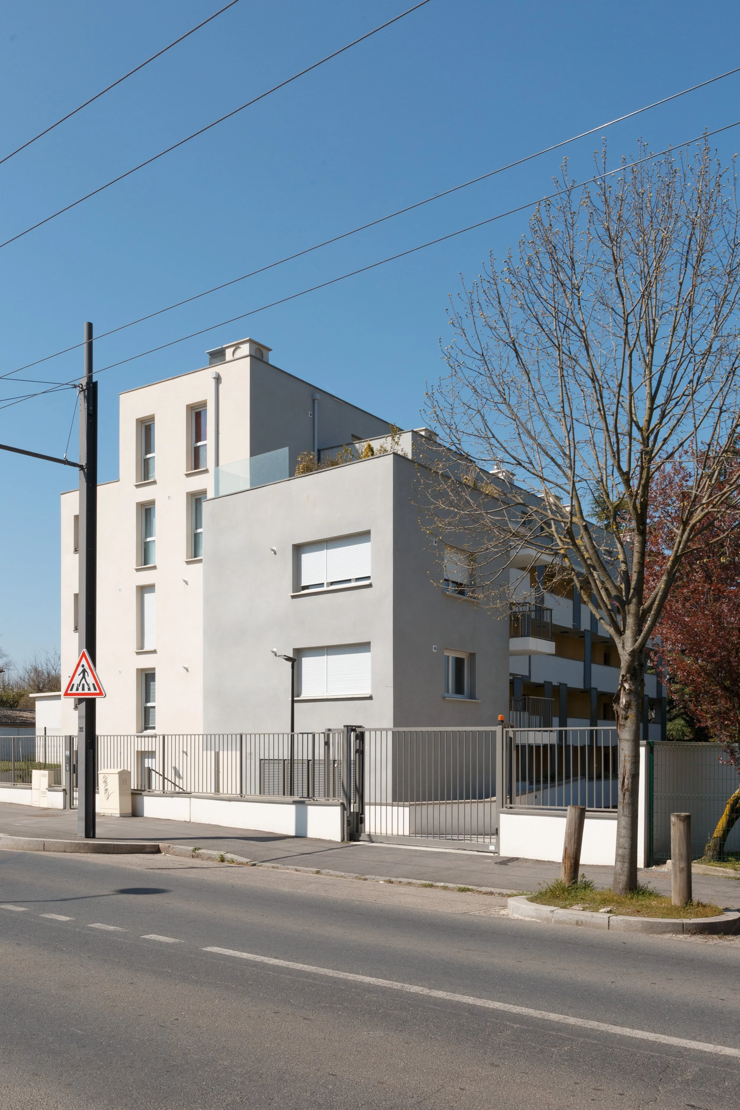 Un bâtiment résidentiel blanc avec plusieurs étages, entouré d'une clôture en métal, avec un arbre nu à proximité. Le ciel est clair et bleu.