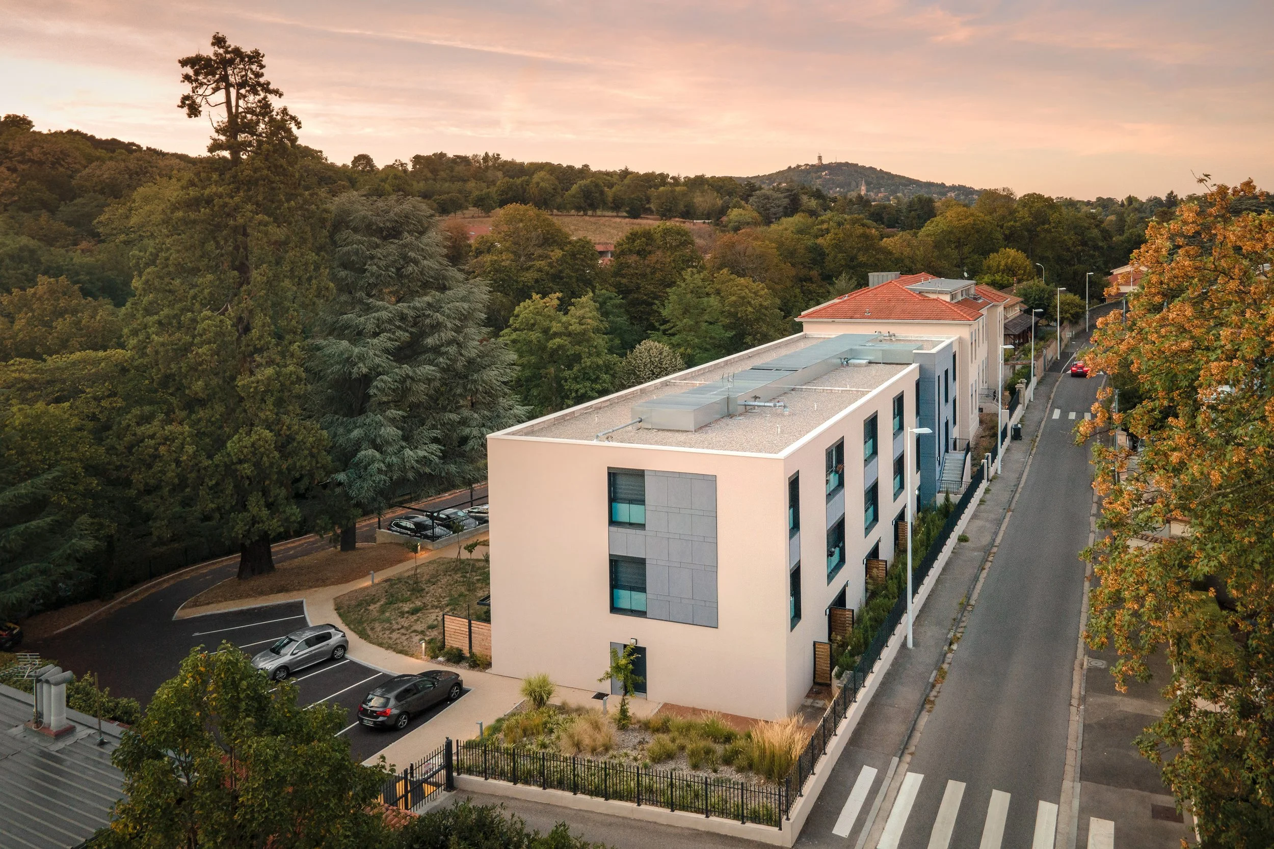 Vue aérienne d'un bâtiment moderne à côté d'une zone boisée en soirée avec un ciel orange.