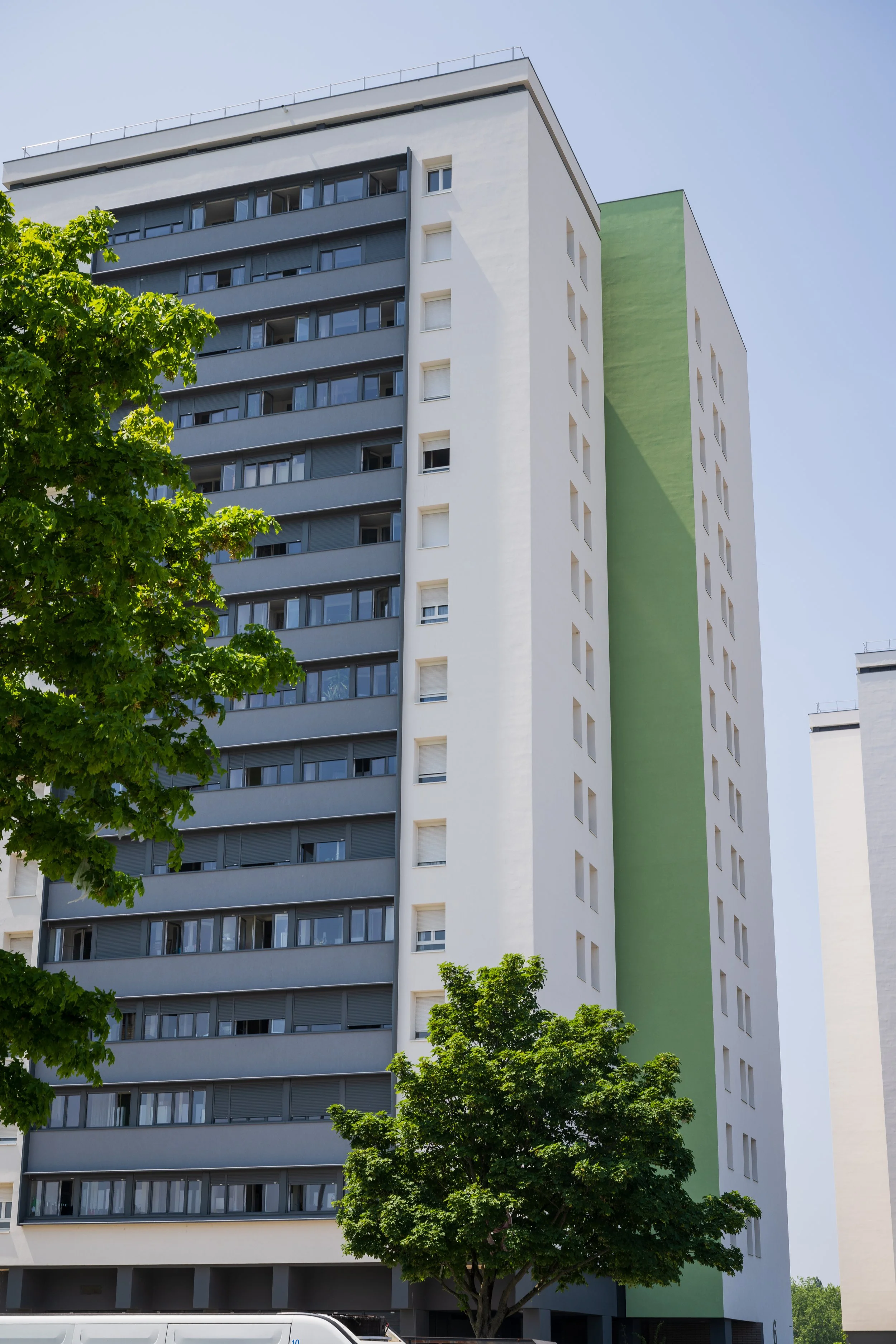 Un immeuble résidentiel moderne avec plusieurs étages, fenêtres et balcons, avec quelques arbres verts devant et un ciel clair en arrière-plan.