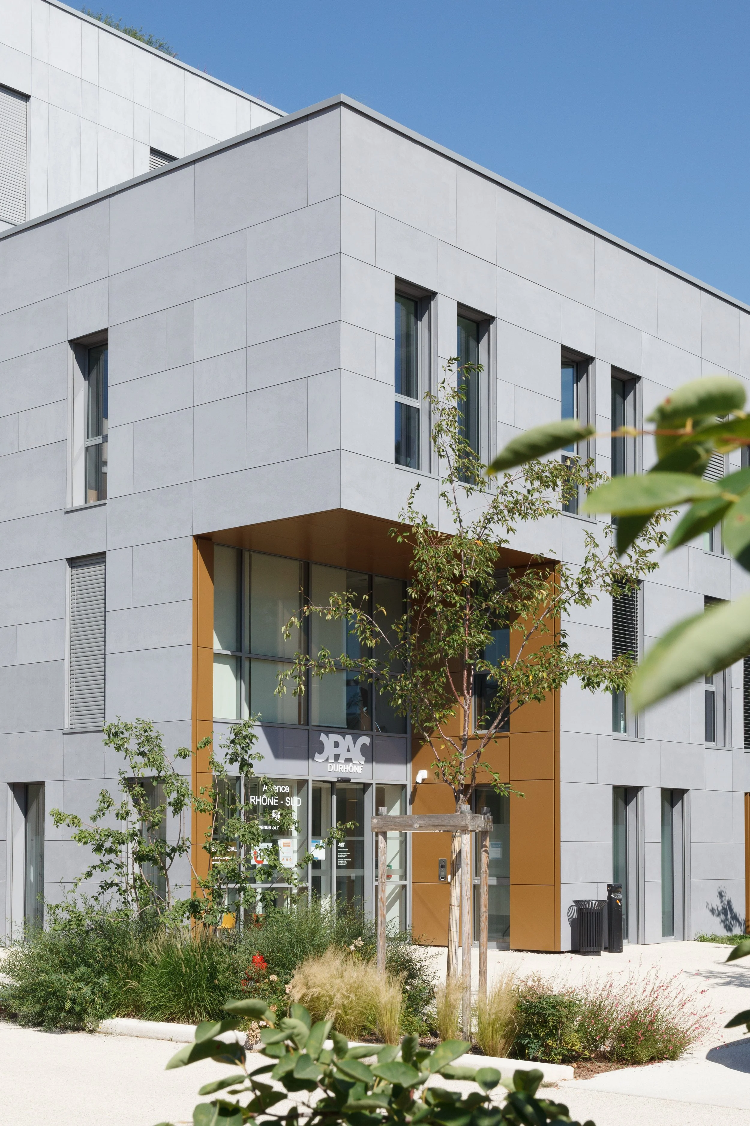 Bâtiment moderne en pierre blanche avec fenêtres rectangulaires, entrée principale avec le logo JPAC Durhône, entouré de végétation et d'arbres, ciel bleu clair.