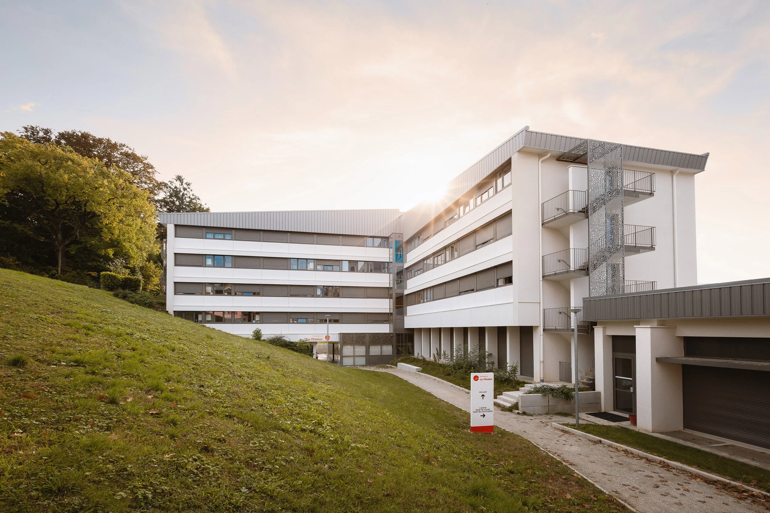 Bâtiment moderne avec plusieurs étages, balcons et fenêtres, située près d'un espace vert, avec un arbre et un panneau d'information à proximité, sous un ciel en fin d'après-midi.