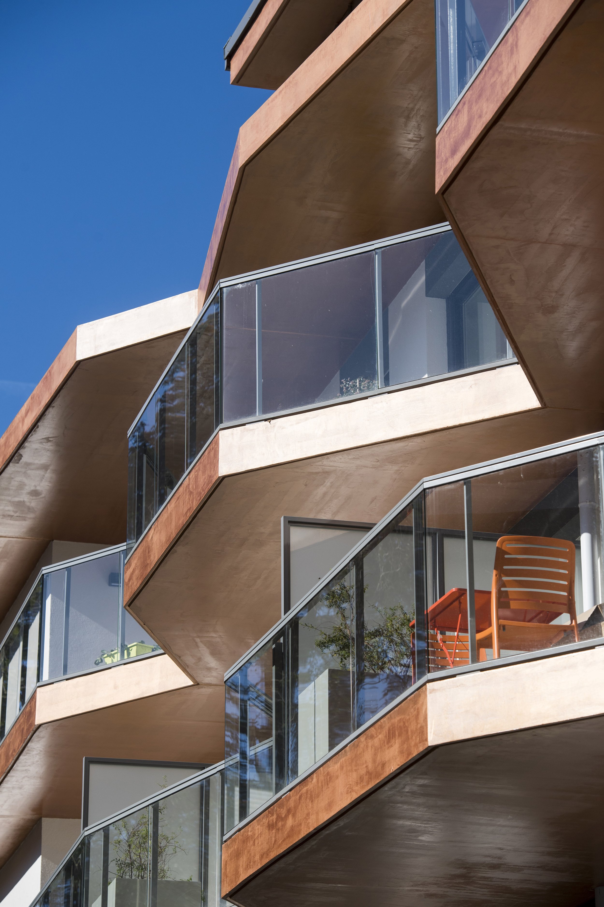 Bâtiment moderne avec balcons en forme d'angle, en façade en béton beige et verre, sous un ciel bleu.
