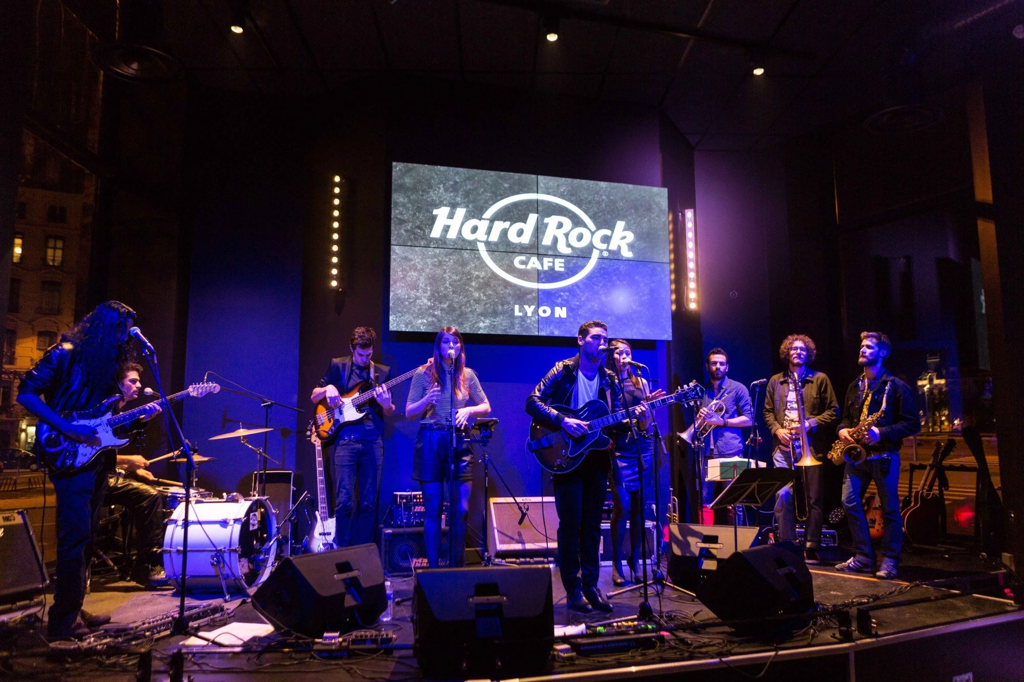 Groupe de musiciens jouant en concert sur scène dans un lieu nommé Hard Rock Café Lyon, avec un grand écran derrière eux affichant le logo du Hard Rock Café.