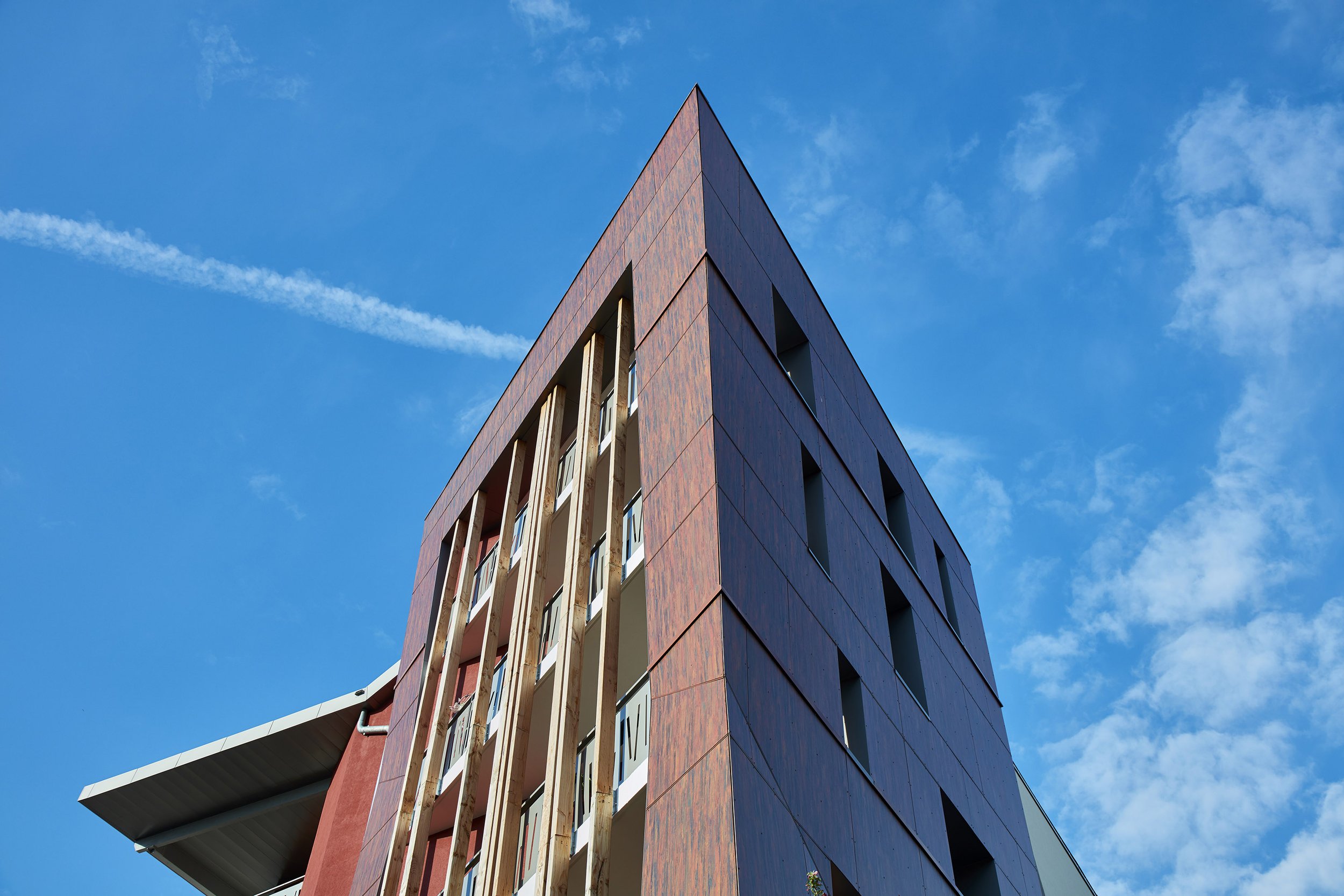 Vue d'un bâtiment moderne à plusieurs étages avec une façade en briques rouges et des fenêtres, sous un ciel bleu avec quelques nuages.