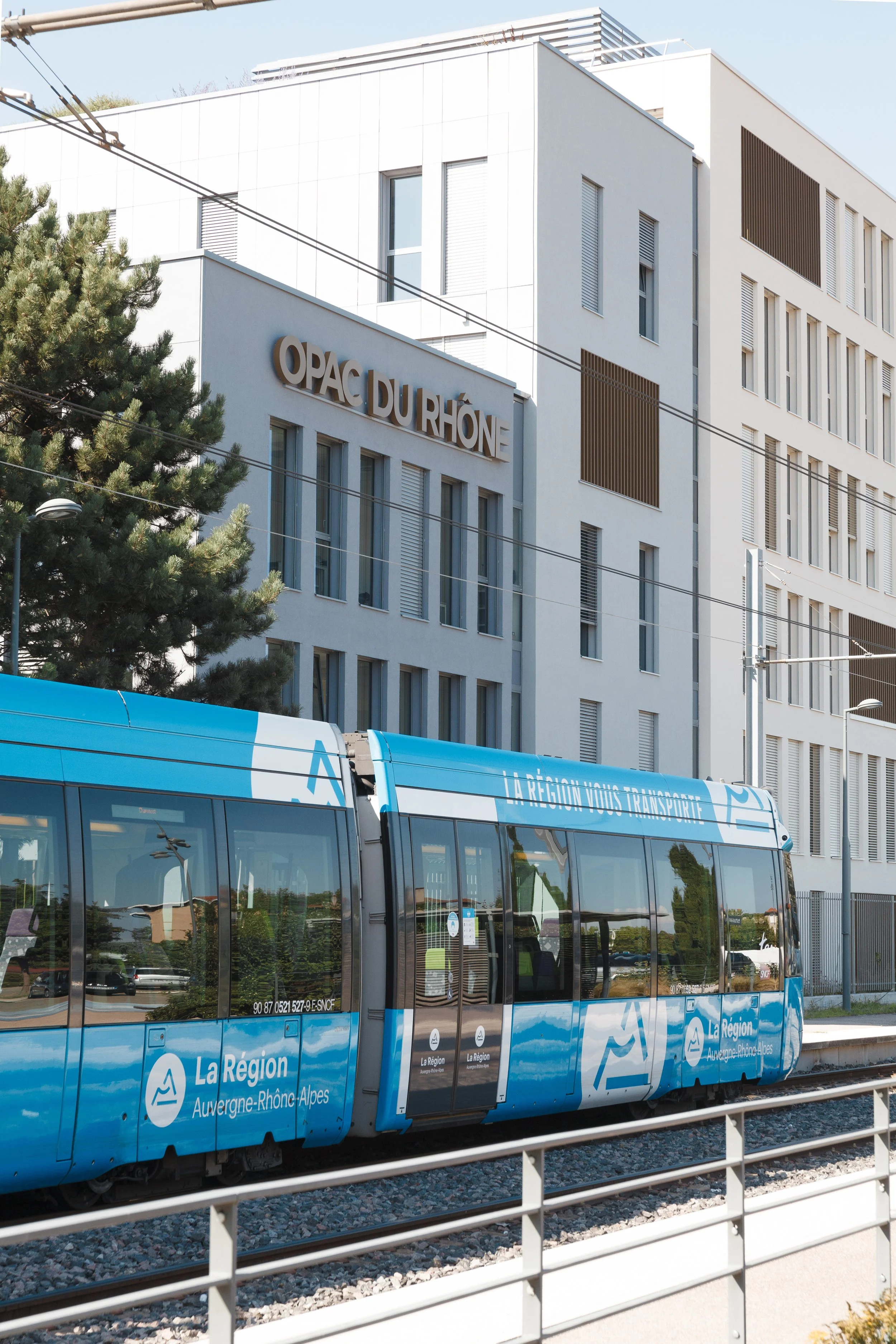 Vue d'un bâtiment blanc moderne avec le texte 'OPAC DU RHÔNE' affiché. En avant-plan, un tramway bleu portant le logo de la région Auvergne-Rhône-Alpes, stationné en gare. Fils électriques suspendus au-dessus du tramway, avec un arbre à gauche.