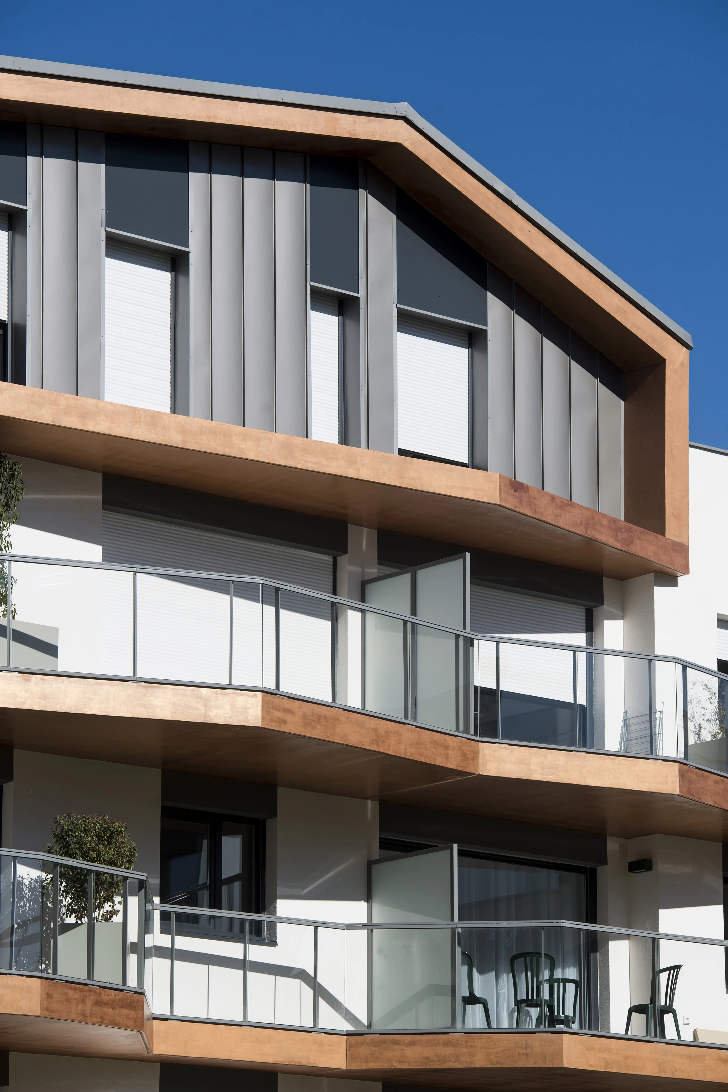 Façade d'un immeuble moderne avec plusieurs balcons en béton et des fenêtres enveloppées de panneaux métalliques gris, sous un ciel bleu clair.