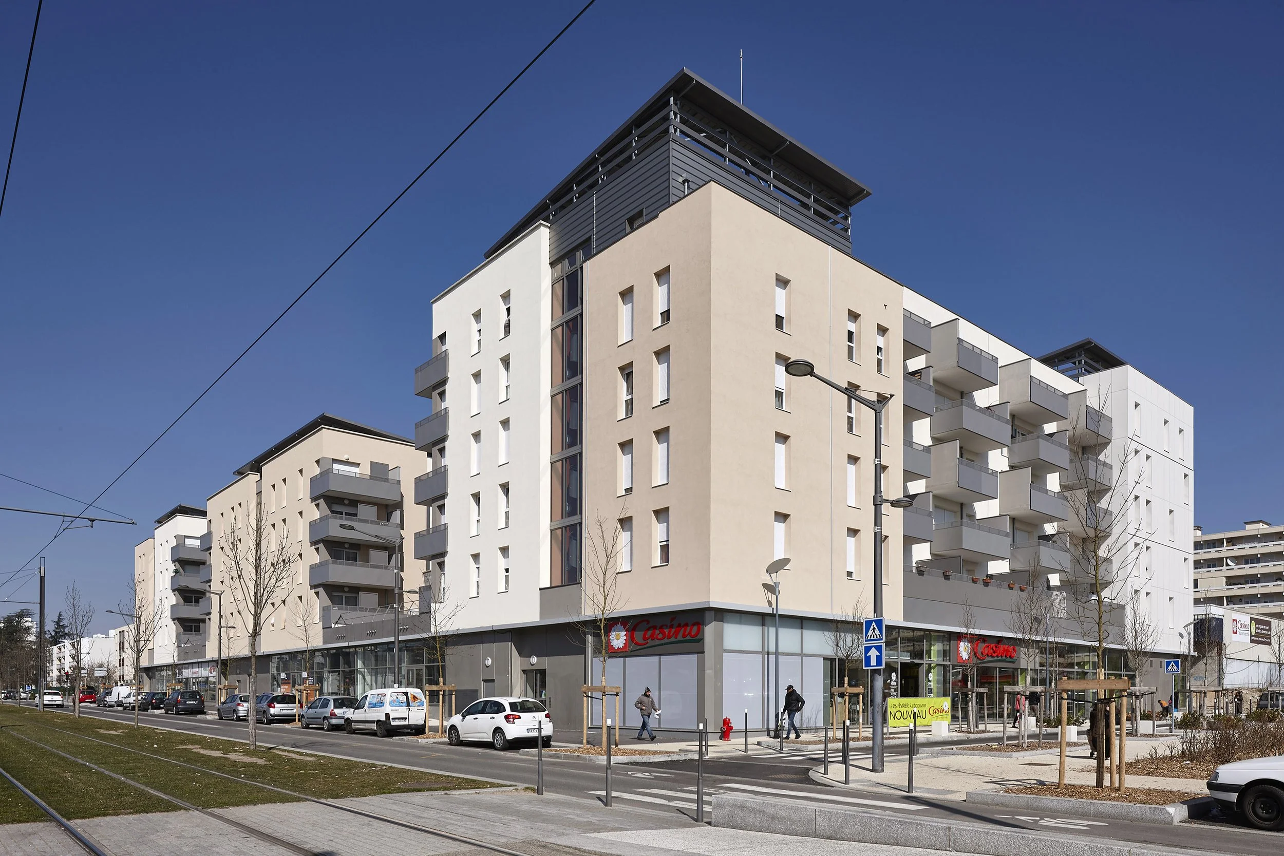 Un immeuble résidentiel moderne avec plusieurs balcons, en bas un magasin avec le signe «Casino», des voitures stationnées, des personnes marchant et des lampadaires. Ciel clair sans nuages.