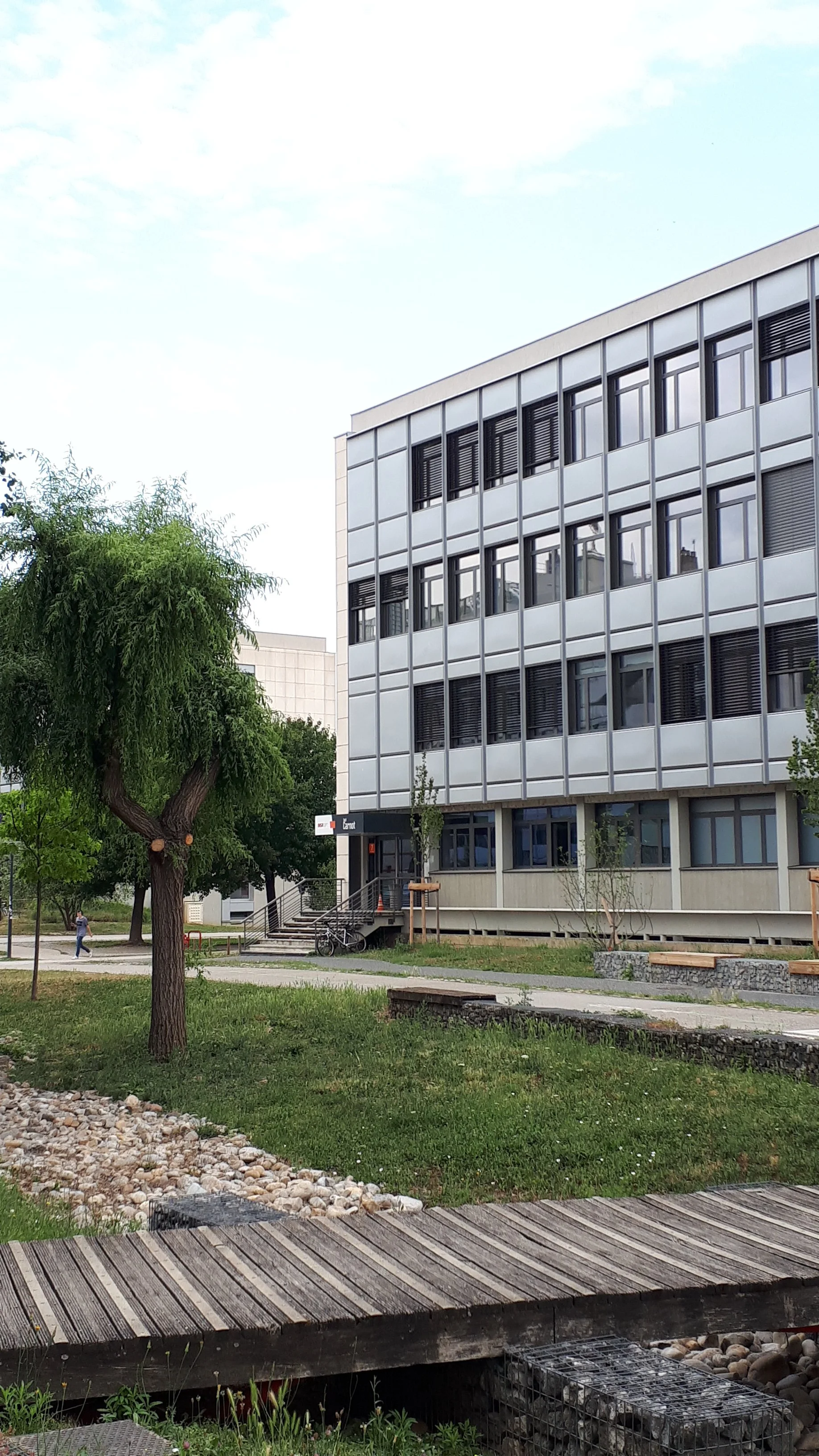Bâtiment moderne en verre et en métal avec un petit parc devant, arbres, un sentier, une personne marchant, un vélo, et un pont en bois dans un environnement urbain.