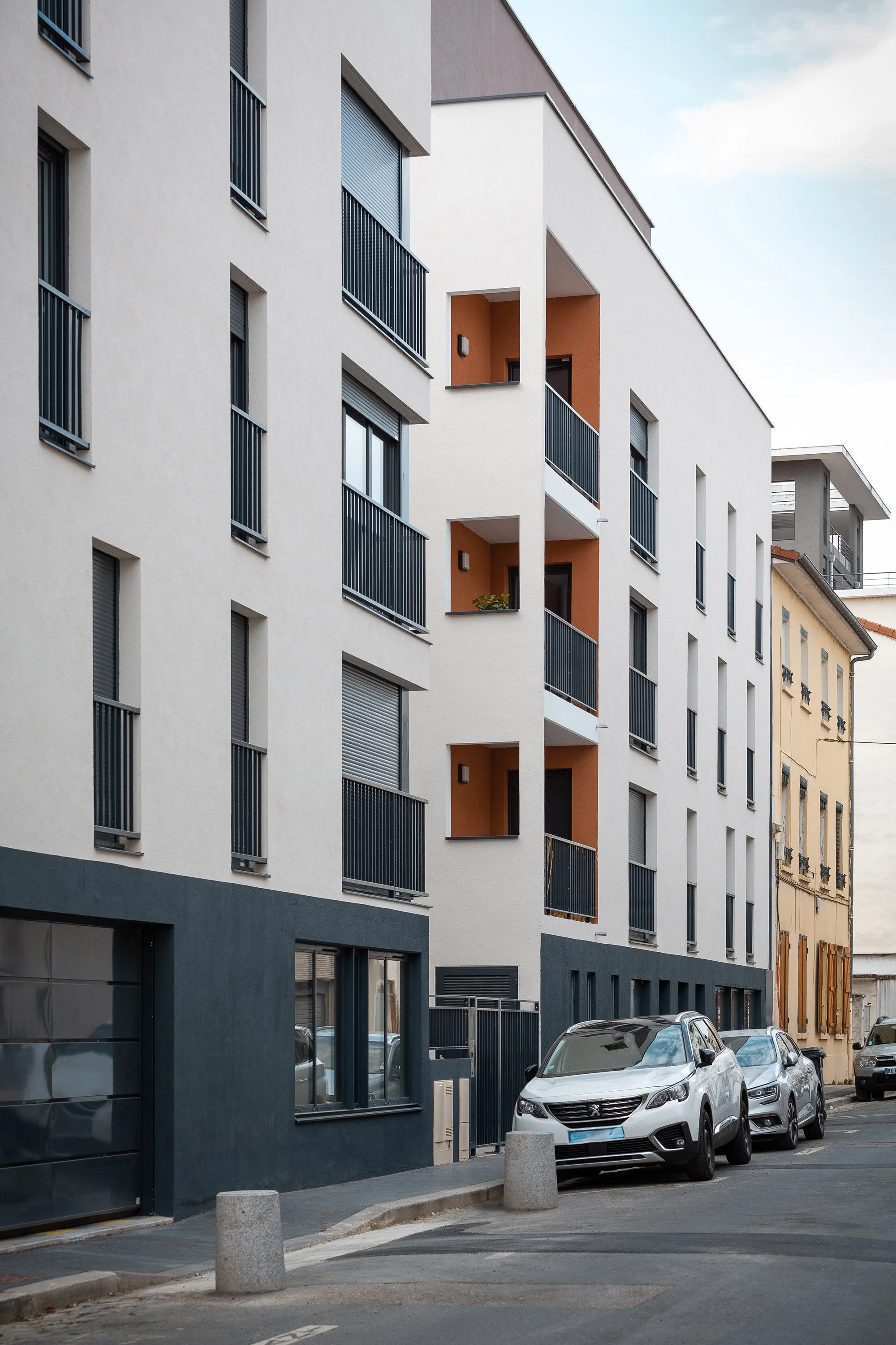 Immeuble moderne avec des balcons en métal noir, voitures garées devant, et ciel partiellement nuageux.