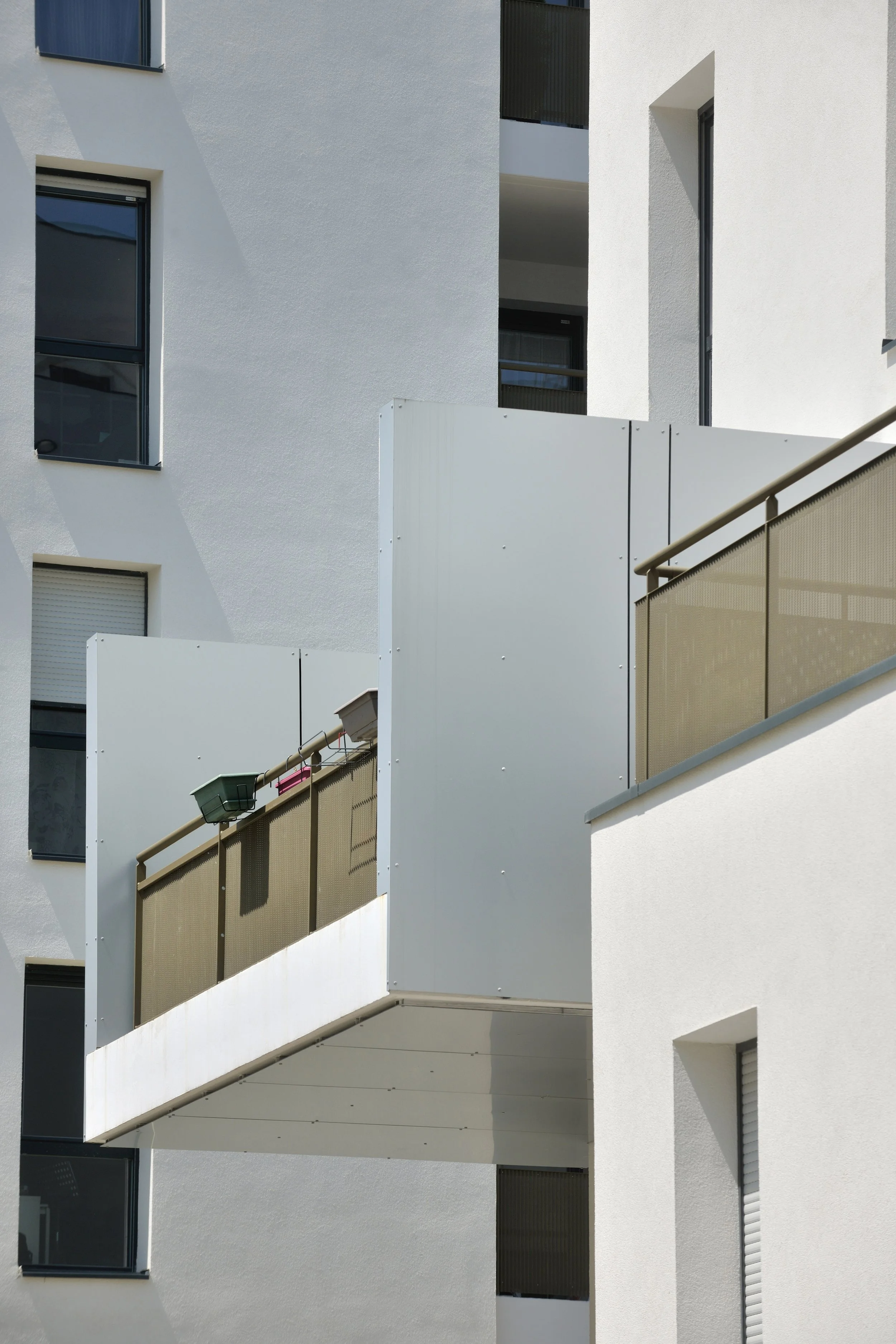 Vue d'un bâtiment résidentiel moderne avec plusieurs fenêtres et balcons avec des portes-fenêtres. La façade est blanche avec des détails en gris. Il y a des balustrades en métal et un volet roulant fermé sur une fenêtre.