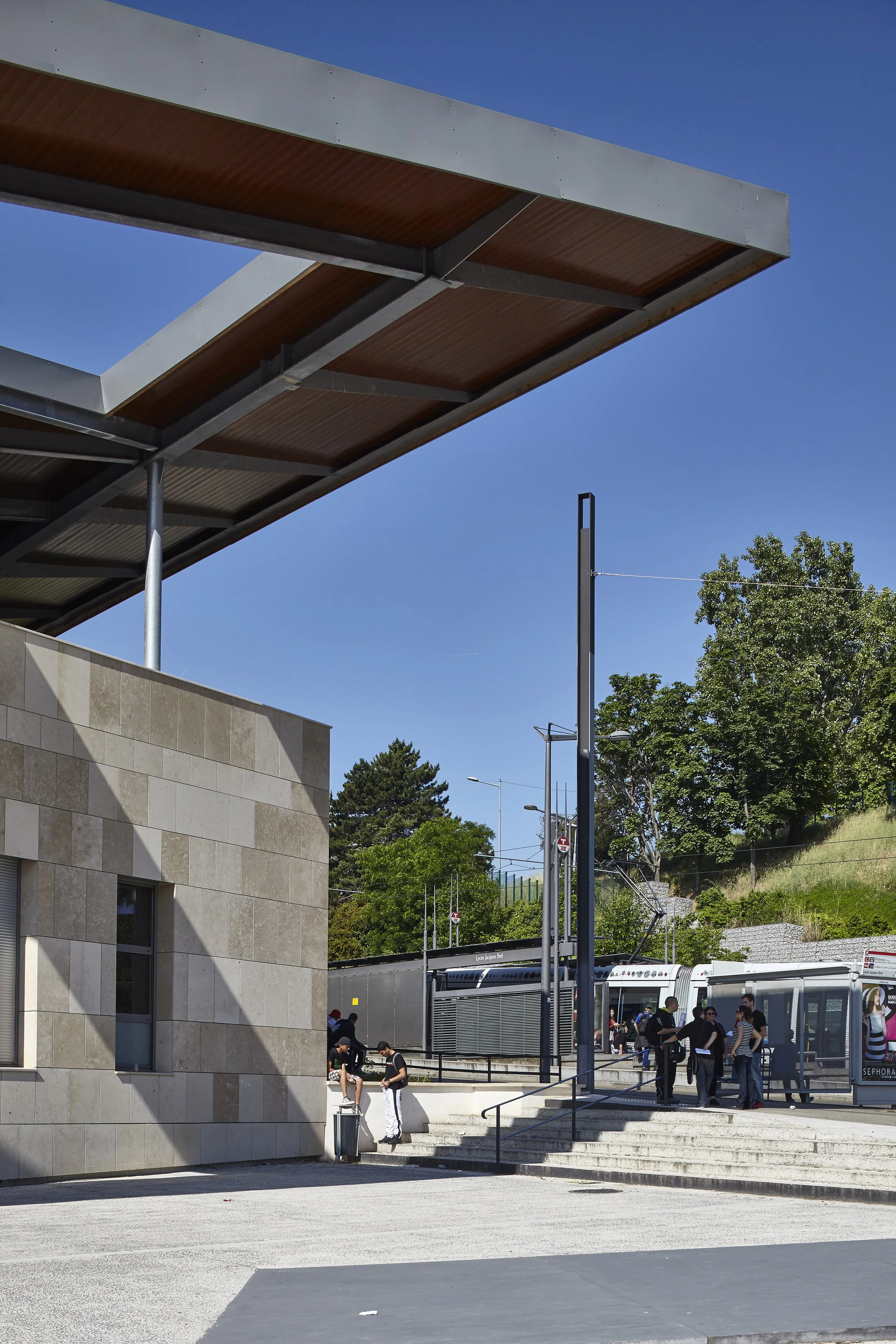 Une station de tramway avec un groupe de personnes en attente. Construite avec des murs en pierre et un toit moderne en métal et bois. Arbres et montagnes en arrière-plan, ciel bleu clair.