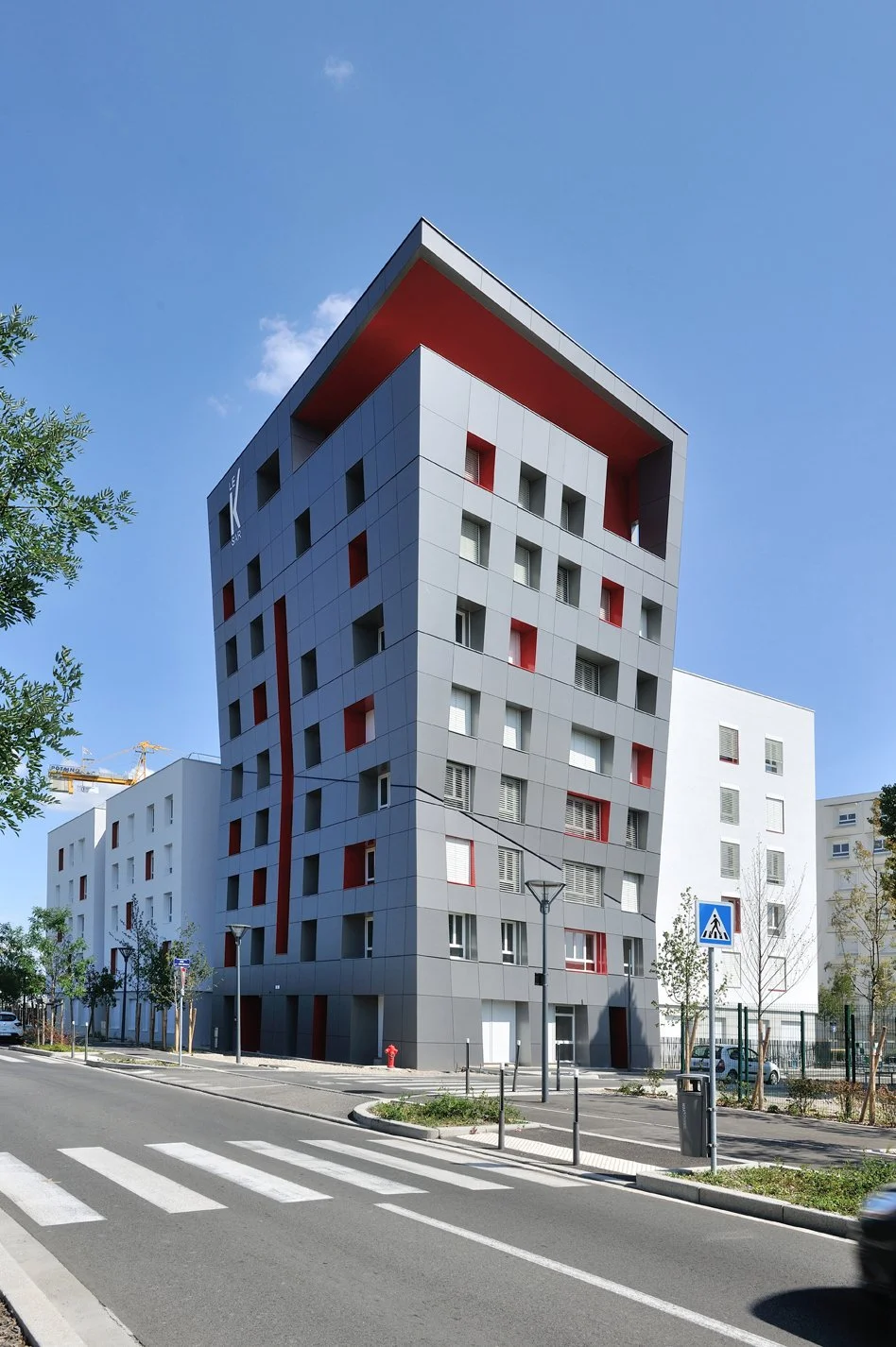 Un bâtiment moderne à plusieurs étages avec une façade en panneaux gris, présentant des fenêtres de différentes tailles et un grand encadré rouge. Le bâtiment est situé dans une zone urbaine avec une rue en premier plan, des arbres, un panneau piéton et un feu de circulation.