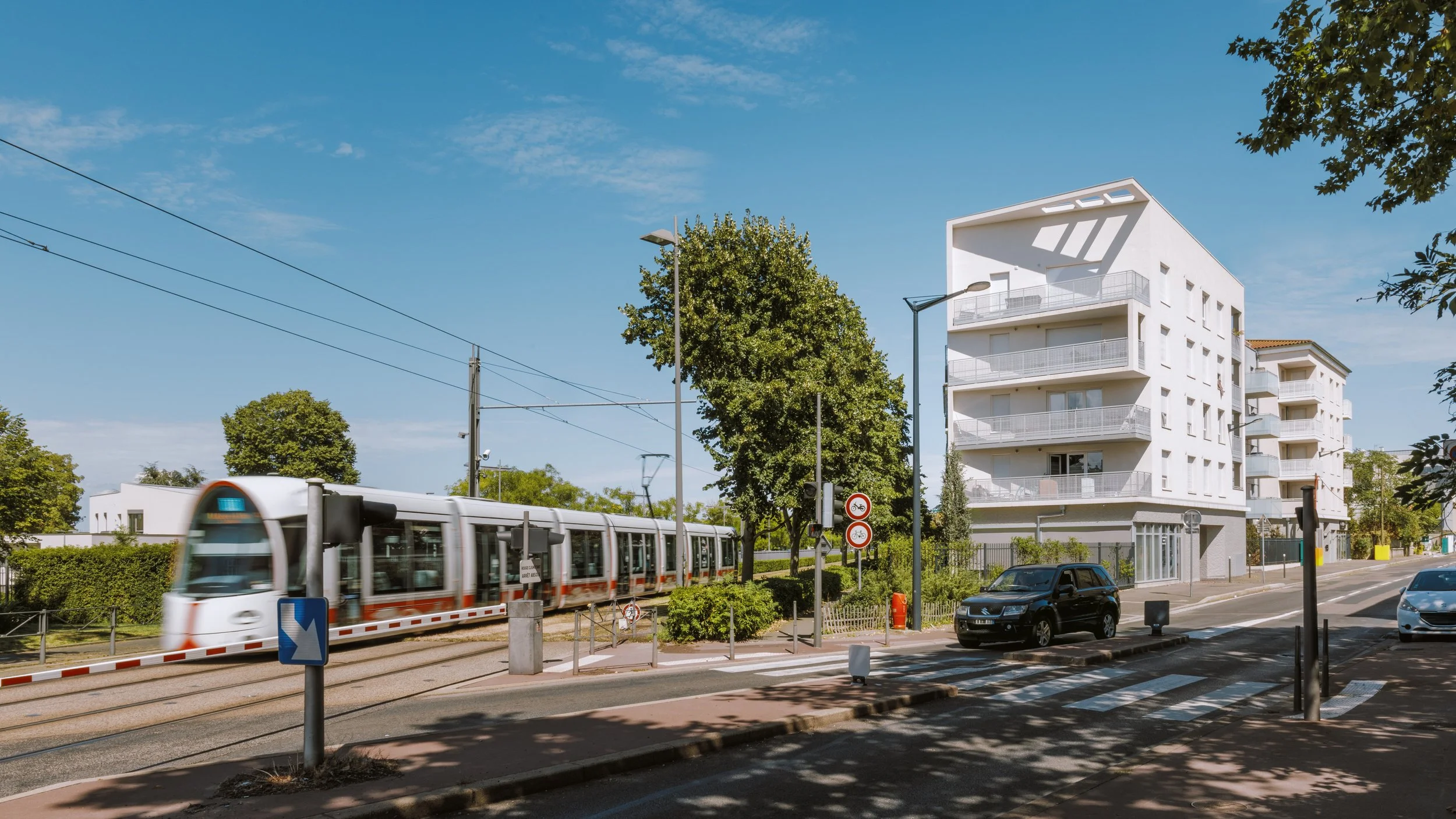 Immeuble moderne blanc avec balcons, tramway en mouvement, arbres et voitures sur la route, ciel clair.