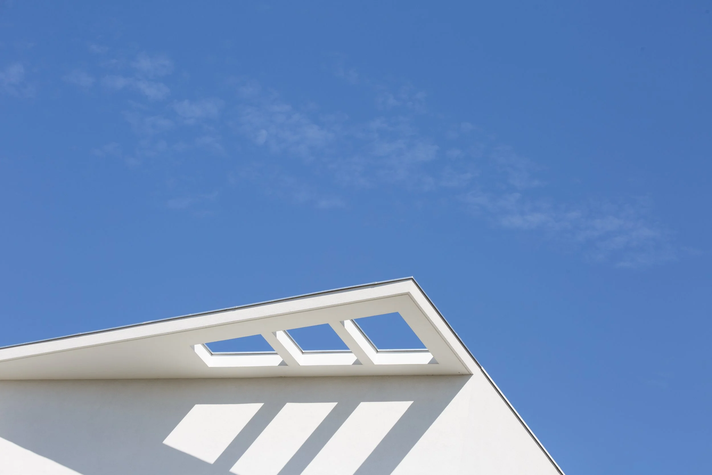 Partie d'une maison moderne en blanc avec une structure en balcons, sous un ciel bleu avec quelques nuages.
