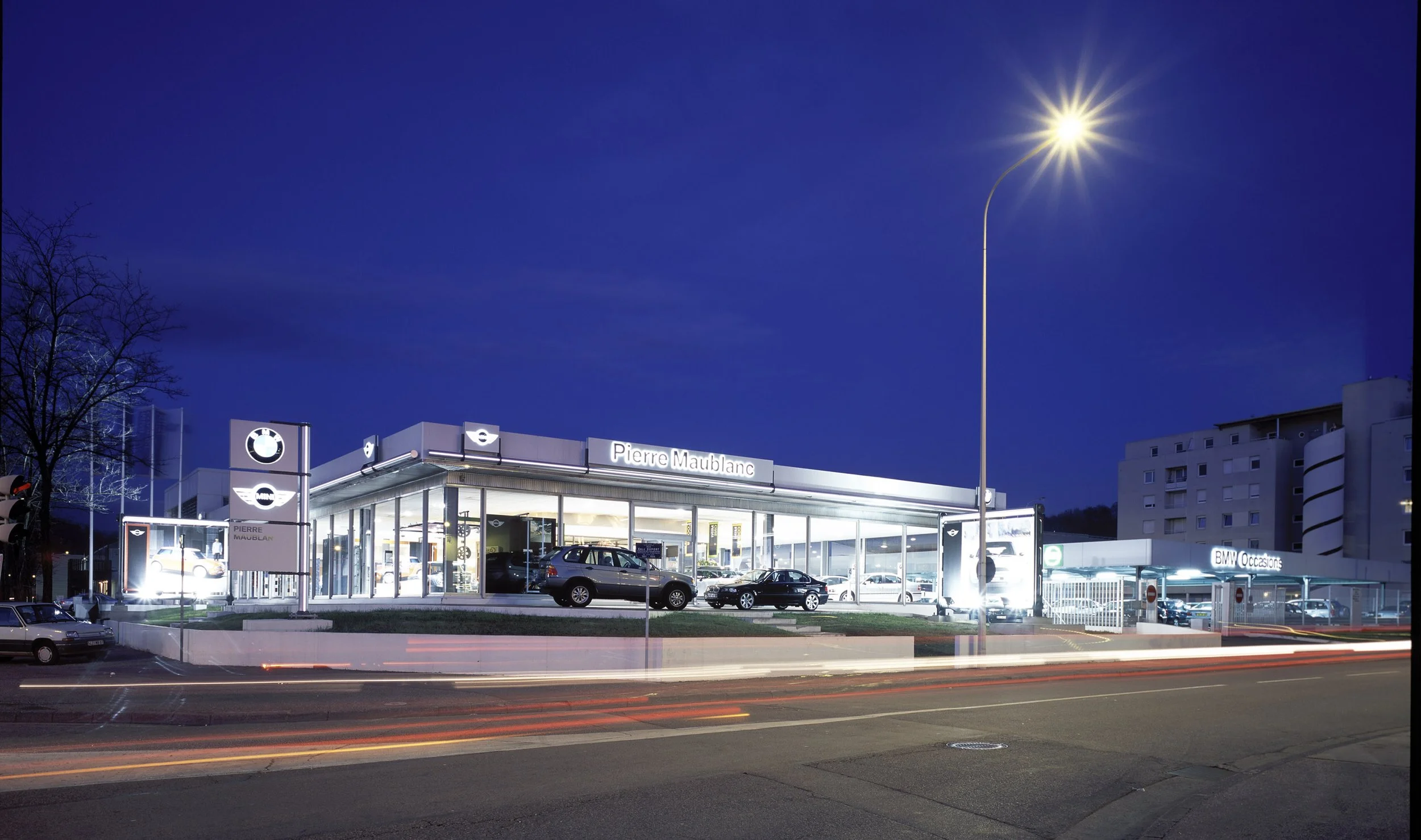 Concessionnaire automobile BMW et Mini, nommée Pierre Maublanc, avec des voitures exposées à l'intérieur et à l'extérieur, au crépuscule, sous un ciel bleu foncé, avec un lampadaire brillant en premier plan.