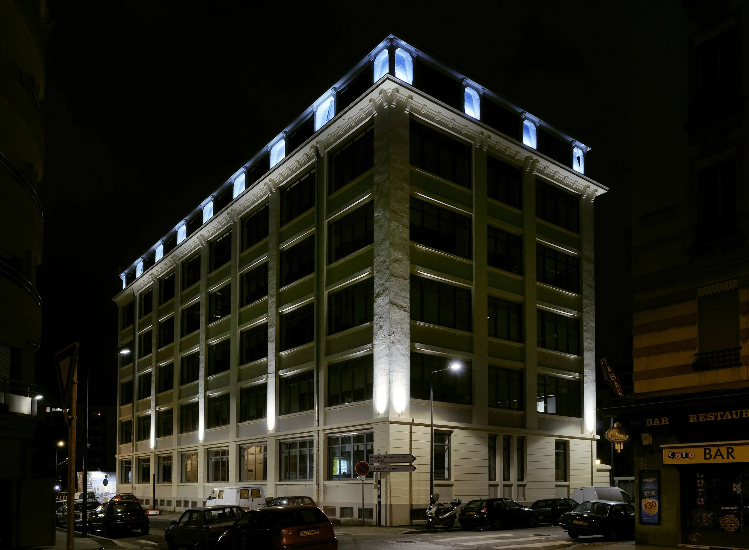 Bâtiment moderne éclairé la nuit, avec plusieurs étages, fenêtres et une façade architecturale élégante, entouré de voitures et de scooters dans la rue.