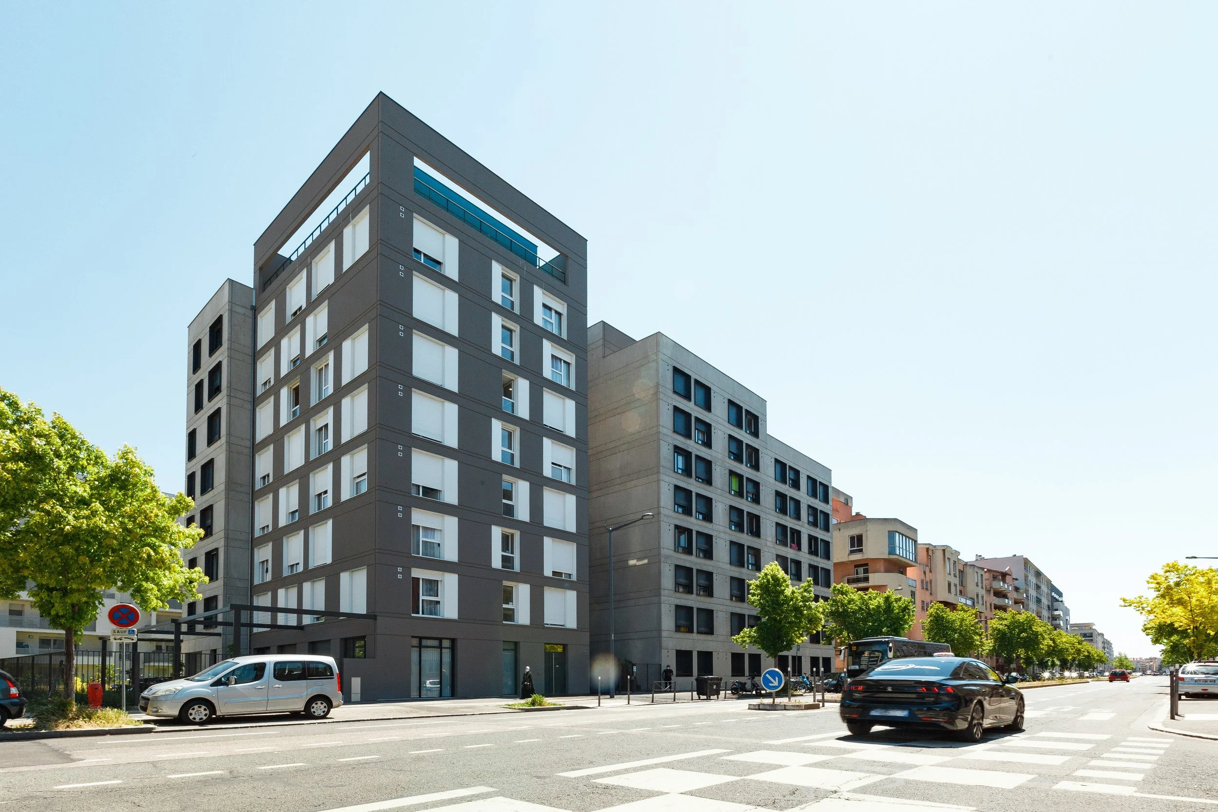 Immeuble résidentiel moderne avec plusieurs étages, fenêtres et balcons, dans une zone urbaine ensoleillée avec des voitures stationnées et des arbres verts en premier plan.
