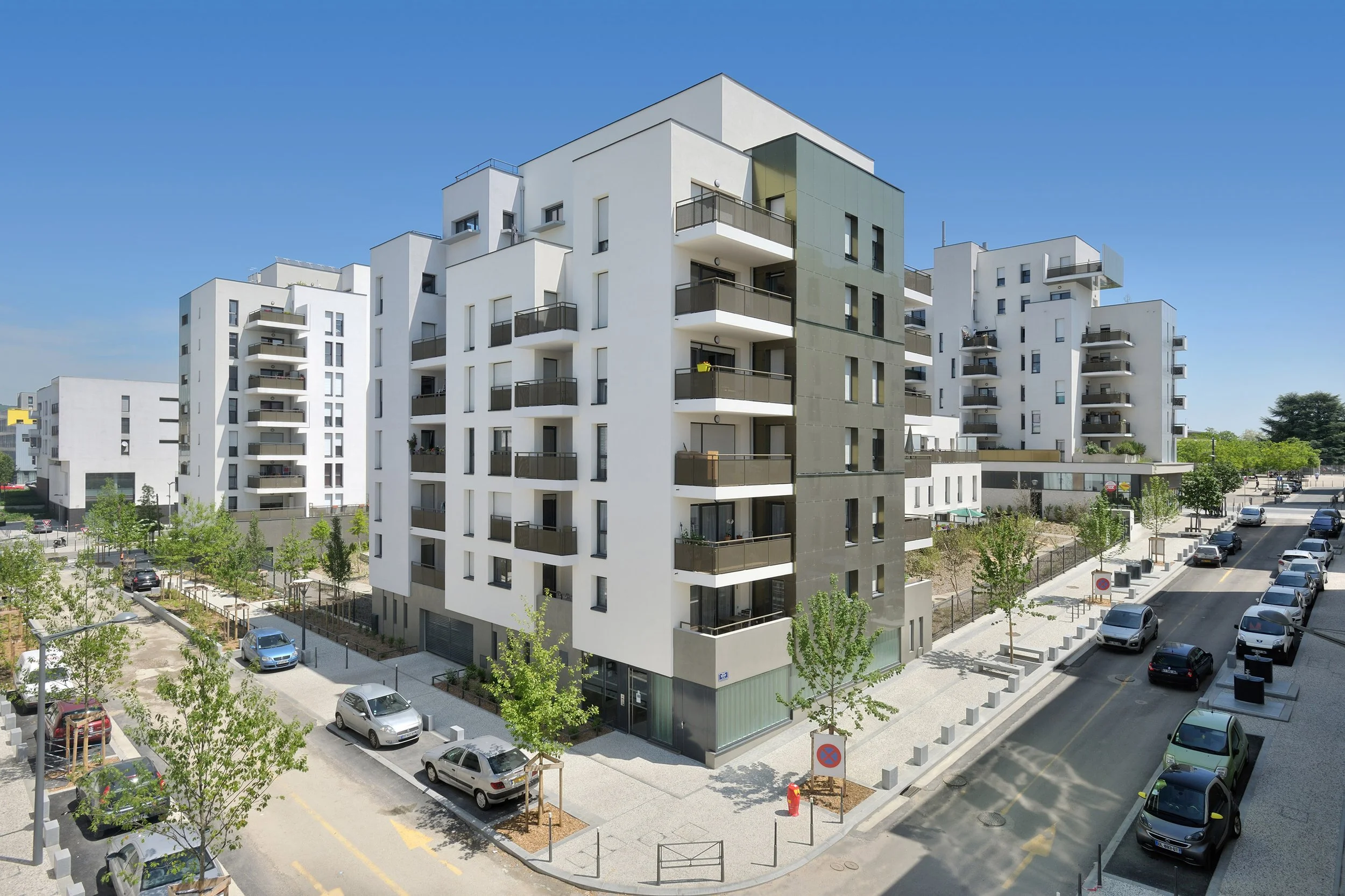 Immeuble résidentiel moderne avec des balcons, vue de face avec parking et arbres en premier plan, ciel bleu clair.