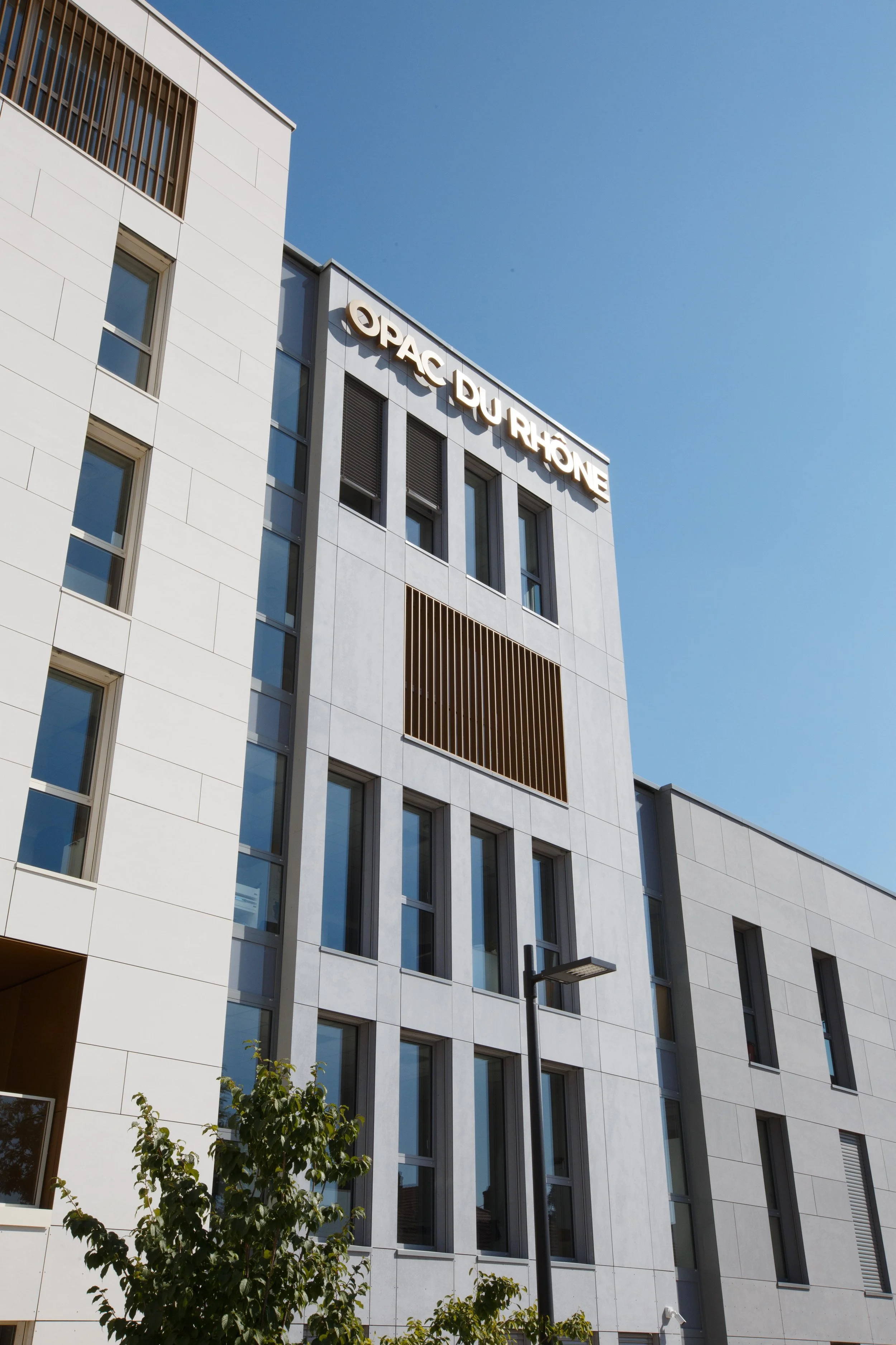 Une façade moderne d'un bâtiment blanc avec plusieurs fenêtres, situé sous un ciel clair et bleu, avec quelques arbres au premier plan.