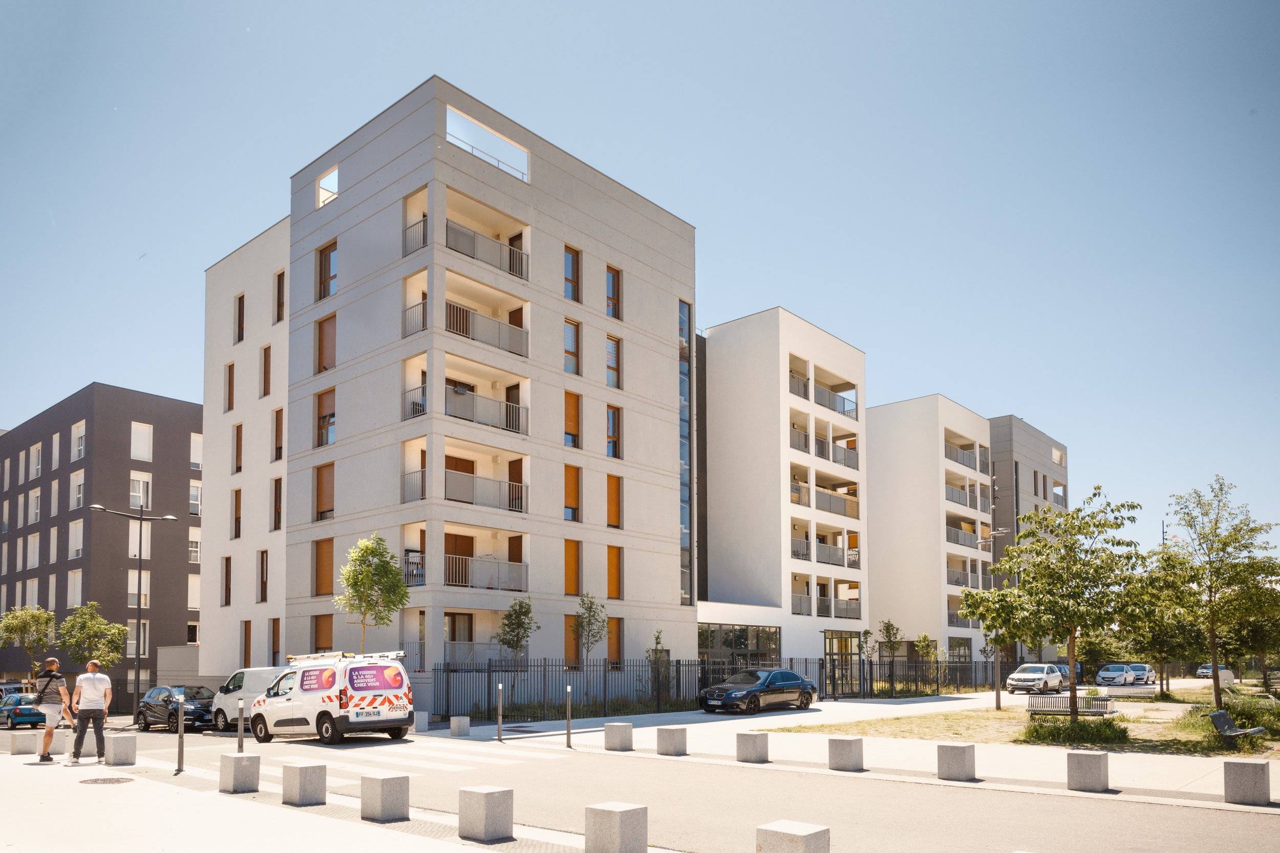 Immeubles résidentiels modernes avec balcons, véhicules garés devant, quelques personnes marchant, arbres et espace public sous un ciel clair.