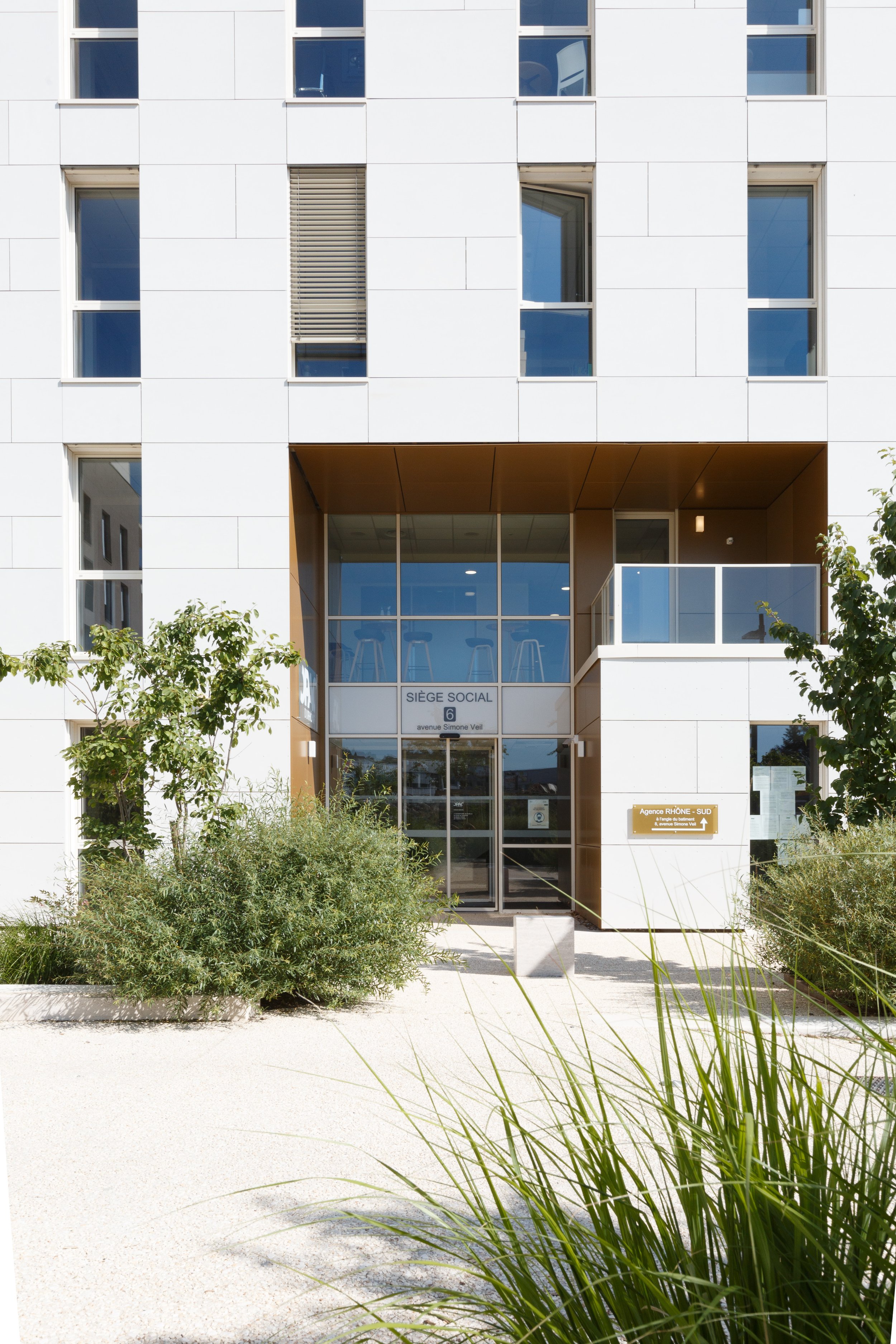 Façade moderne d'un bâtiment blanc avec entrée en verre, signes indiquant un siège social et une agence au sud, entourée de végétation et de gravier.