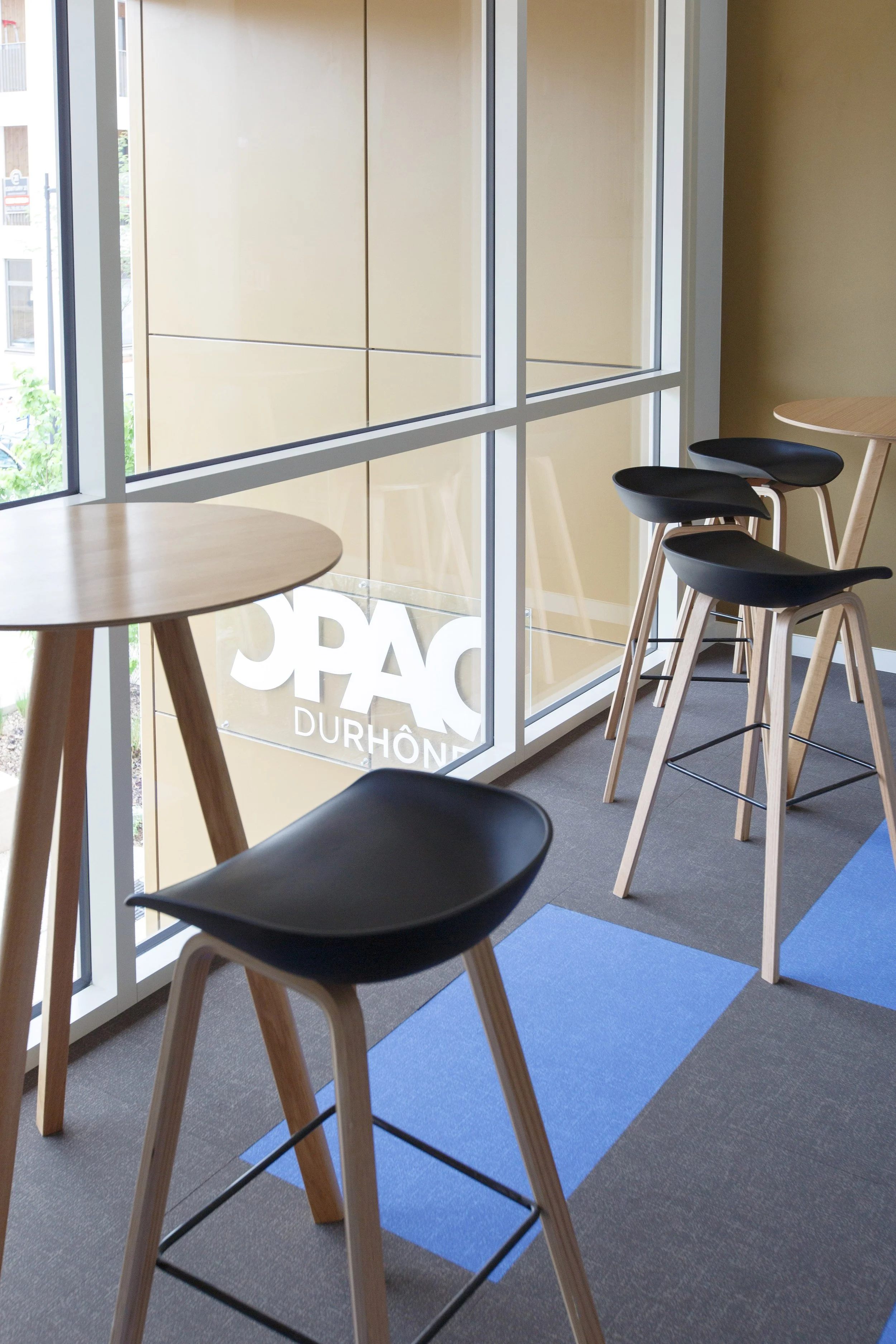 Intérieur d'un café ou restaurant avec des tables en bois et des chaises noires, vue sur une grande fenêtre laissant entrer la lumière naturelle, avec un logo partiel visible à l'extérieur.