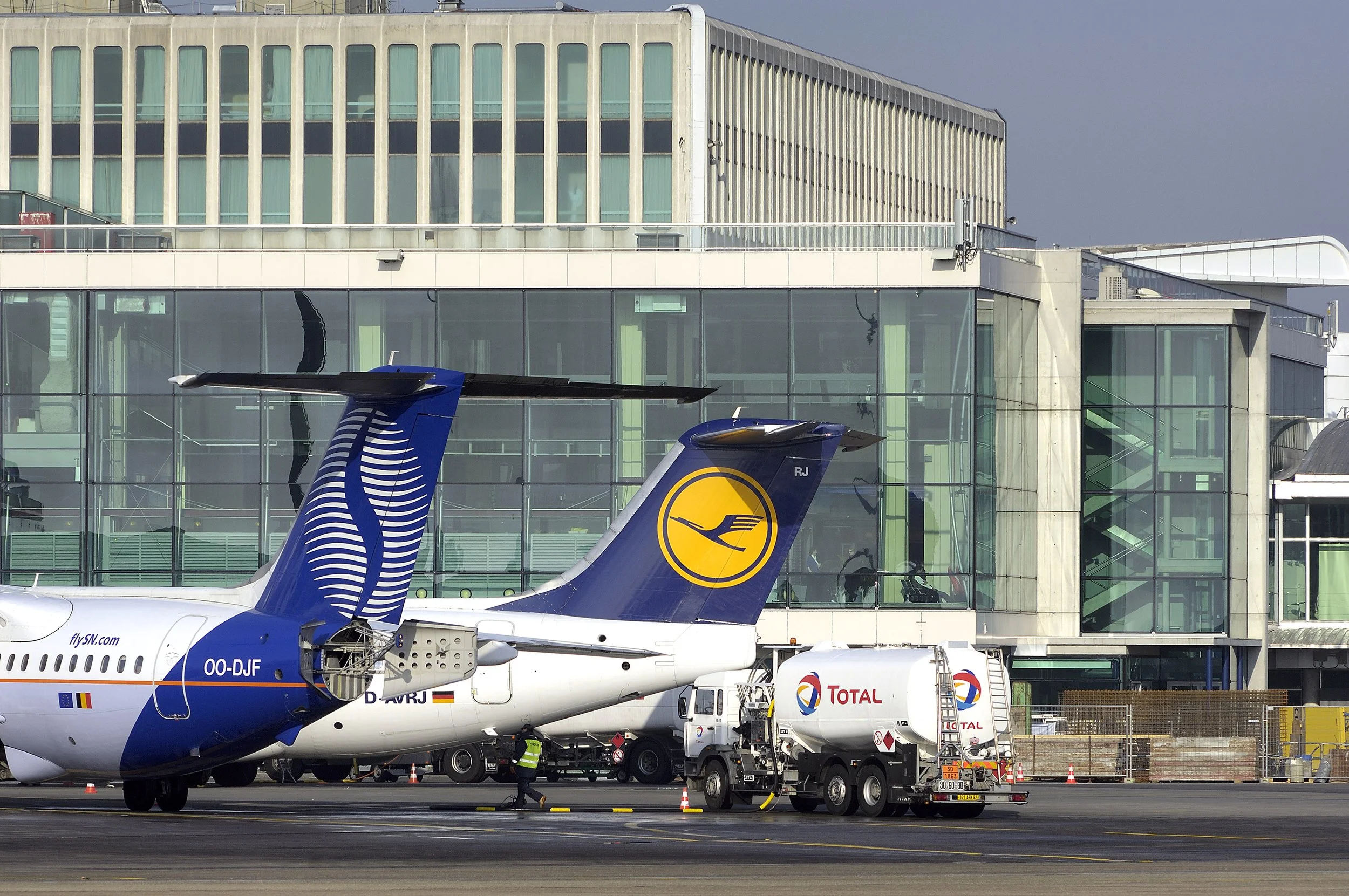 Avion de ligne à l'aéroport avec un terminal en arrière-plan, plusieurs véhicules au sol, et des pompiers ou du personnel de l'aéroport travaillant sur la scène.